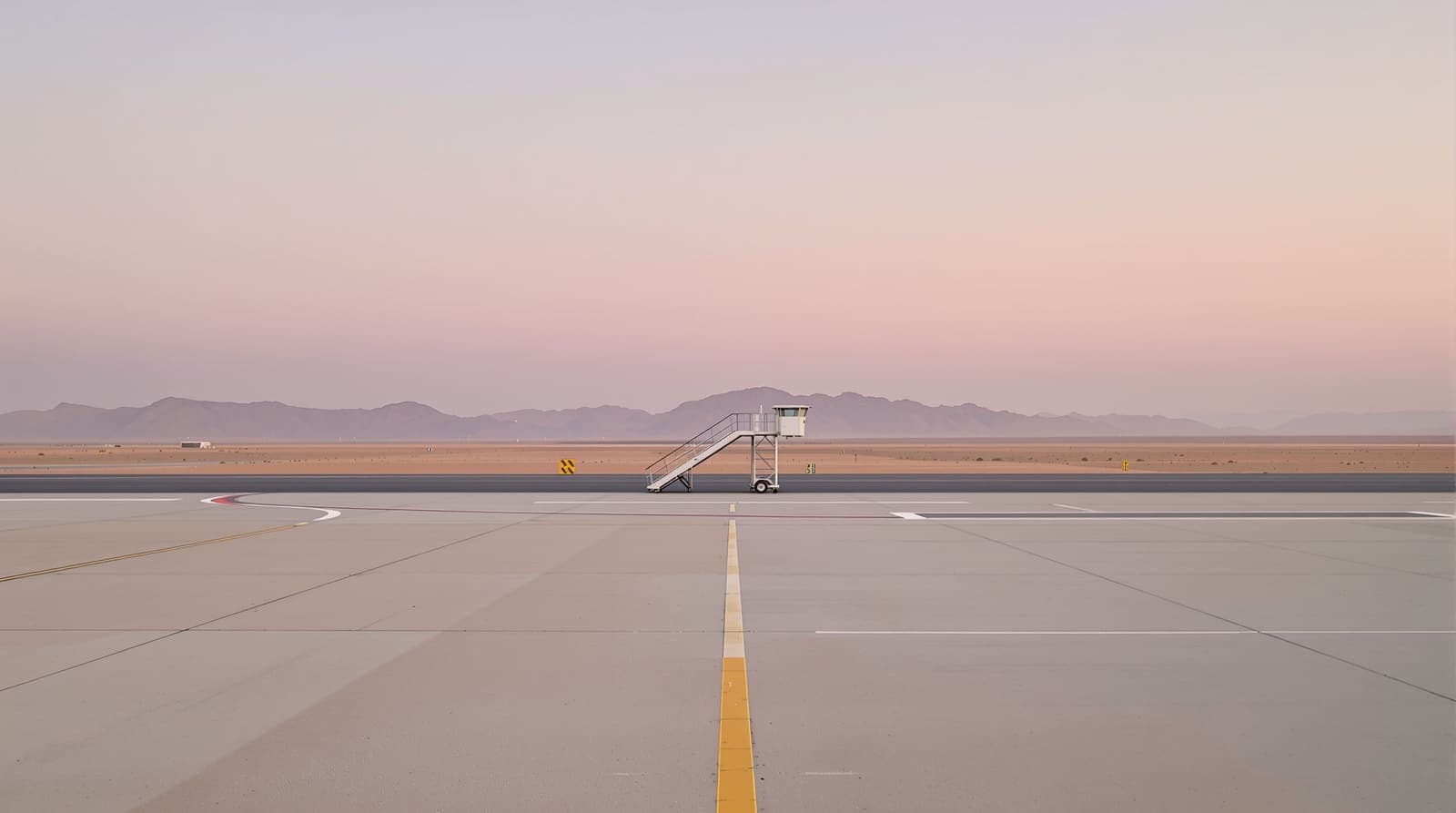 Illustration of an empty desert airport apron at dawn with boarding stairs waiting on the tarmac and rocky ridgelines beyond.