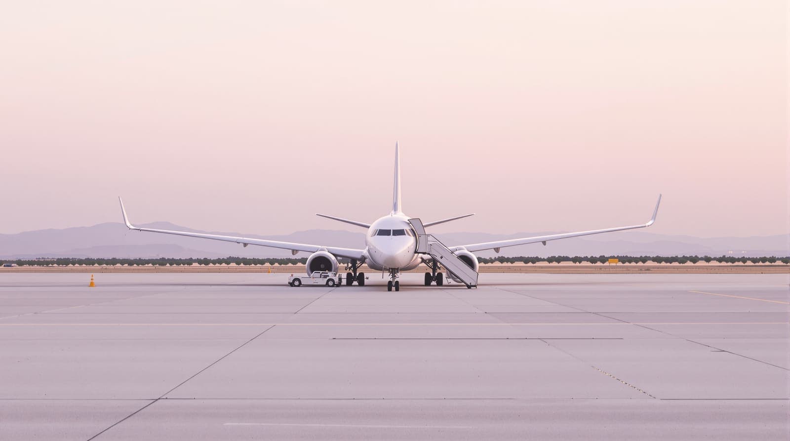 Illustration of a white passenger jet parked on an empty airport apron at dawn, with a stairway at its rear door and low hills beyond.