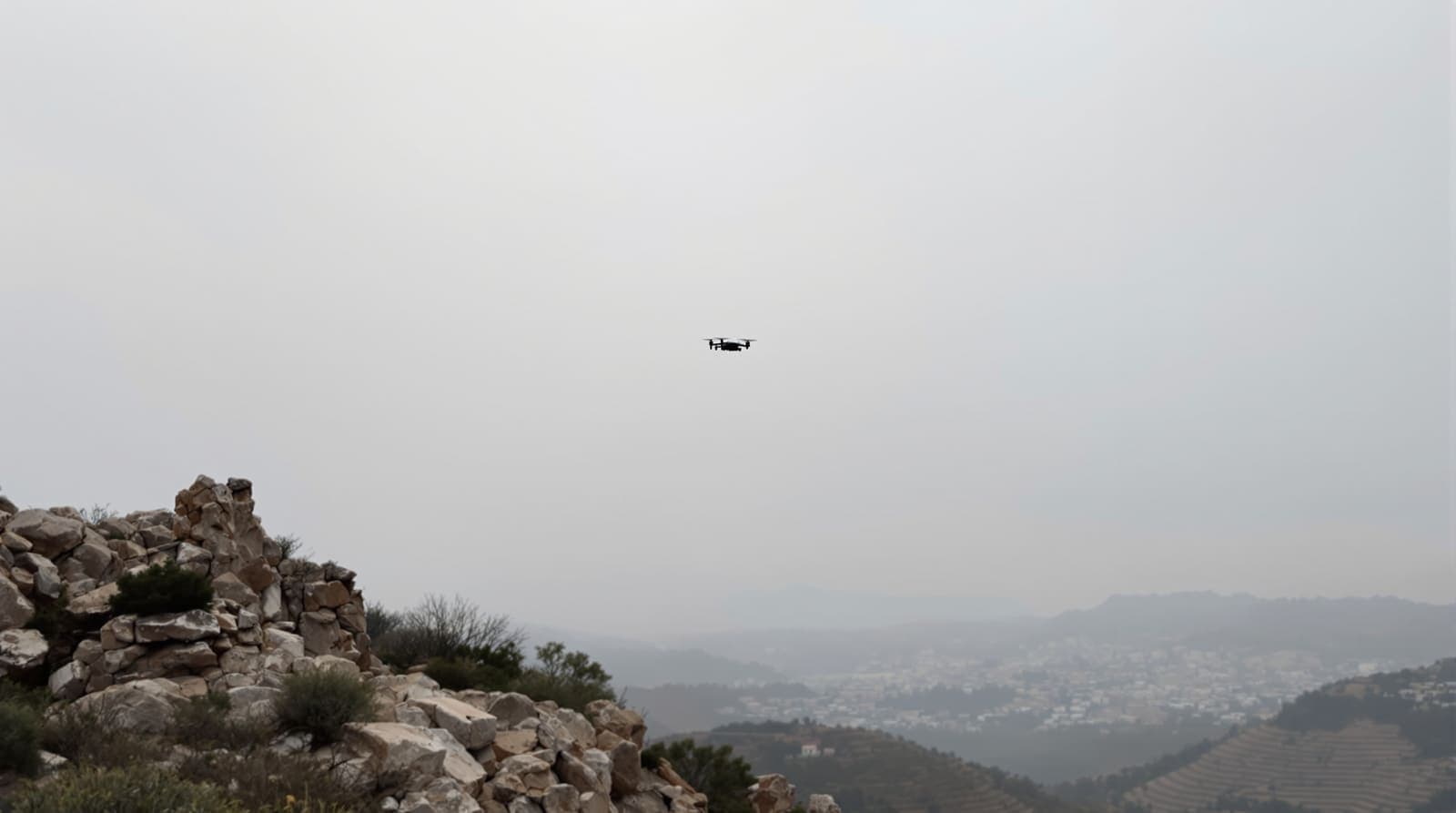 Illustration of a small drone silhouetted high in a pale dawn sky above a rocky scrub ridge in southern Lebanon.