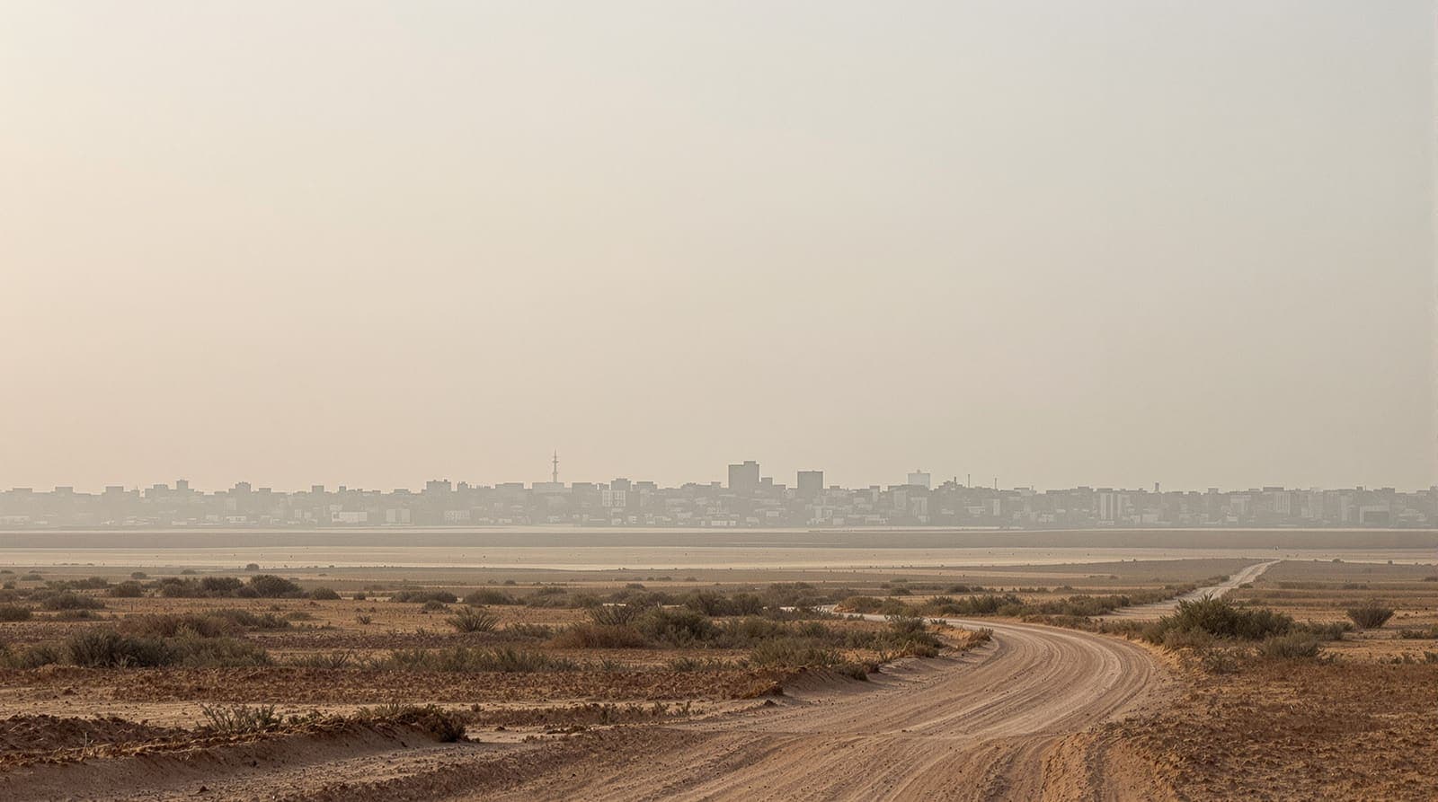 Illustration of the low concrete skyline of Khan Younis in southern Gaza seen across flat sandy farmland under a hazy dawn sky.