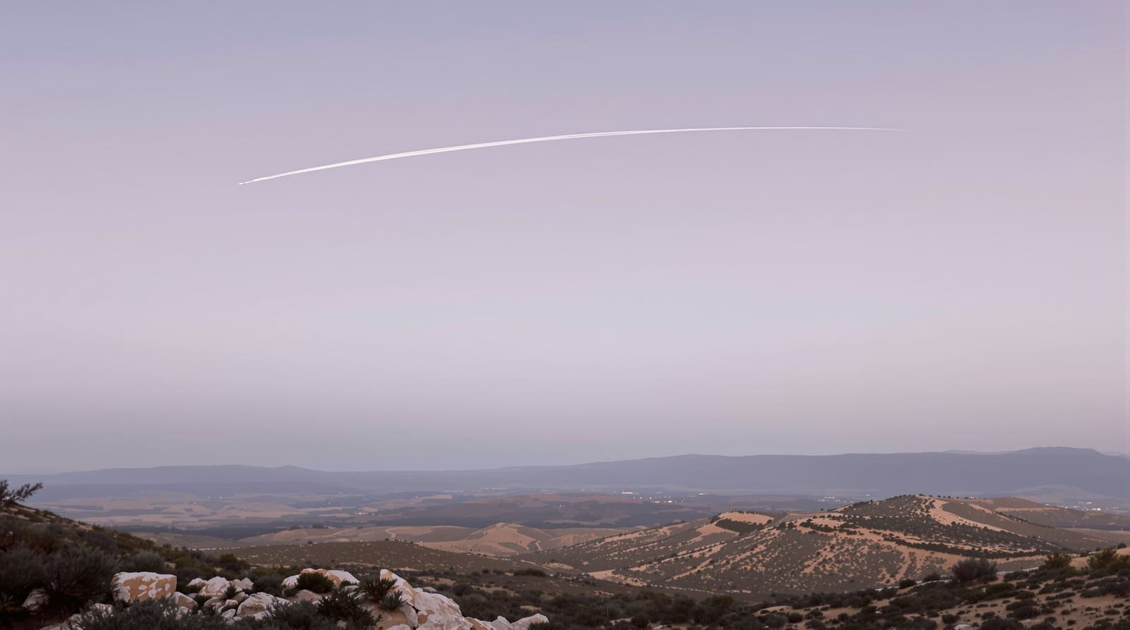 Illustration of a thin white contrail curving across a dusk sky above hazy scrub-covered hills and olive terraces in southern Lebanon.