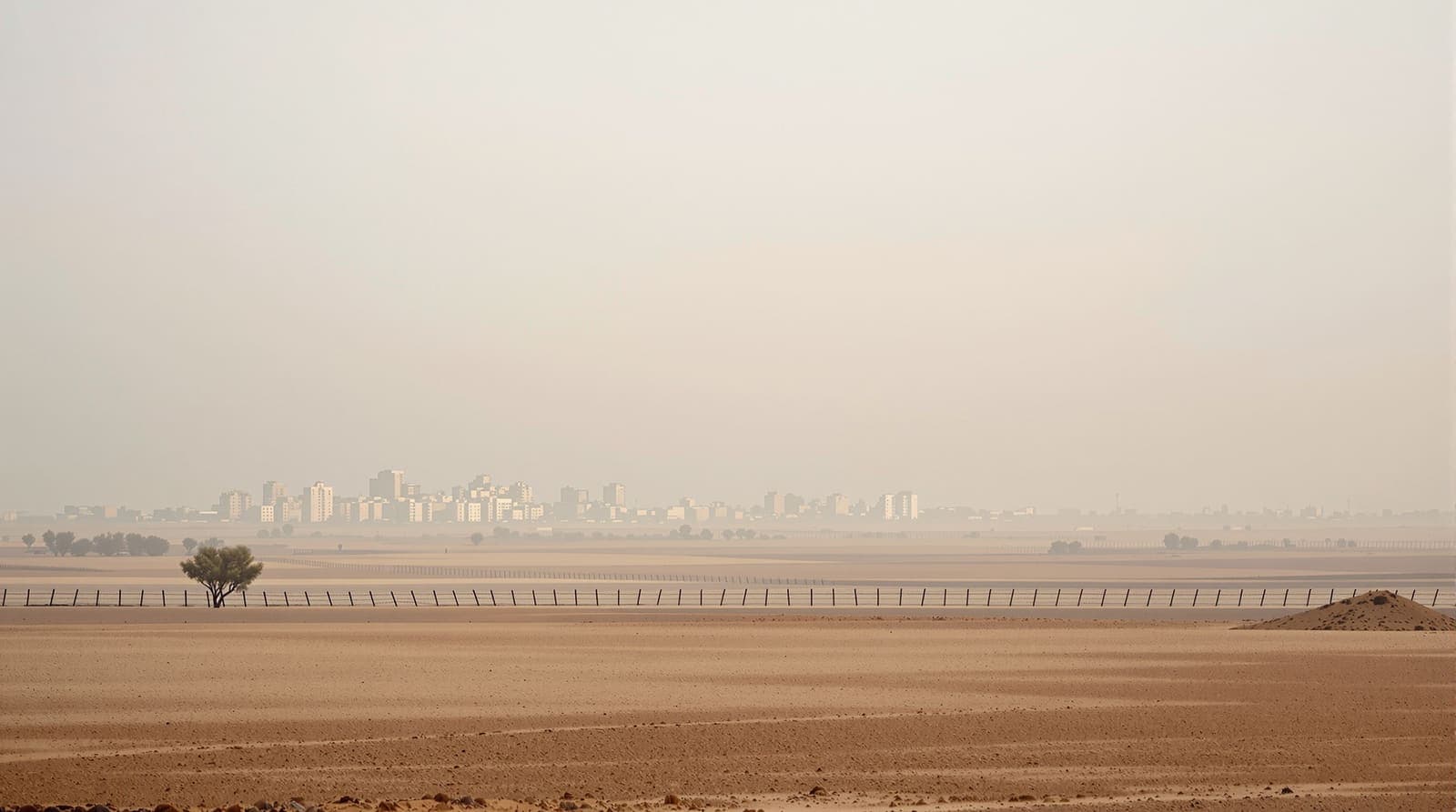 Illustration of the Gaza skyline seen in hazy dawn light from distant farmland beyond a long border fence.