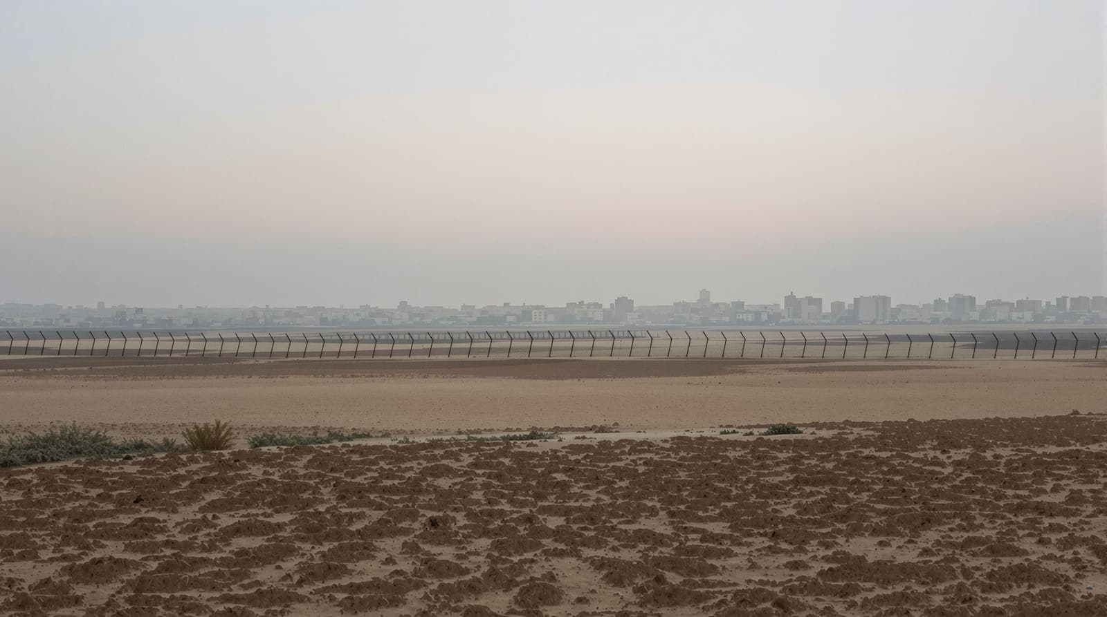 Illustration of the hazy Gaza skyline at dawn seen from across flat farmland and a long border fence.