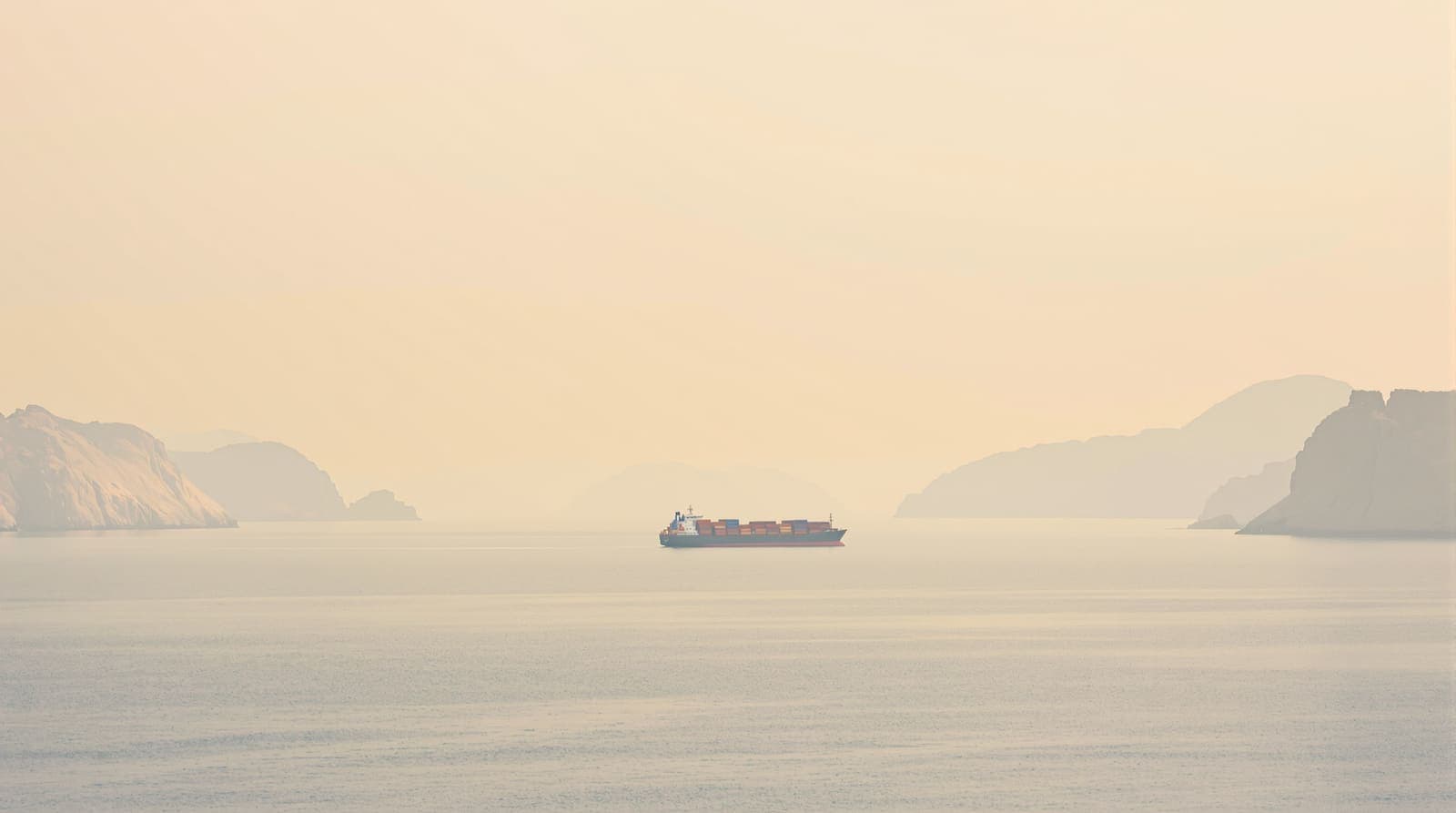 Illustration of a distant container ship on calm water in a narrow strait between hazy arid coastlines at dawn.