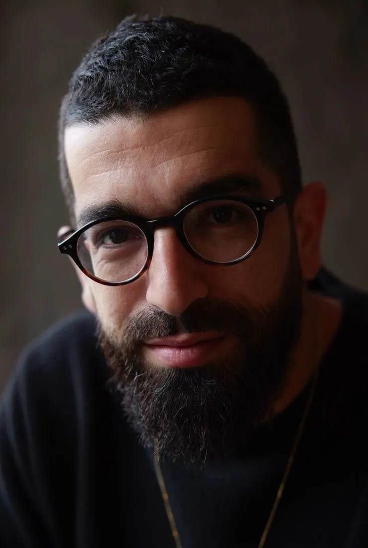 Hanan Ben Ari in round glasses and a dark sweater, a close portrait, 2025