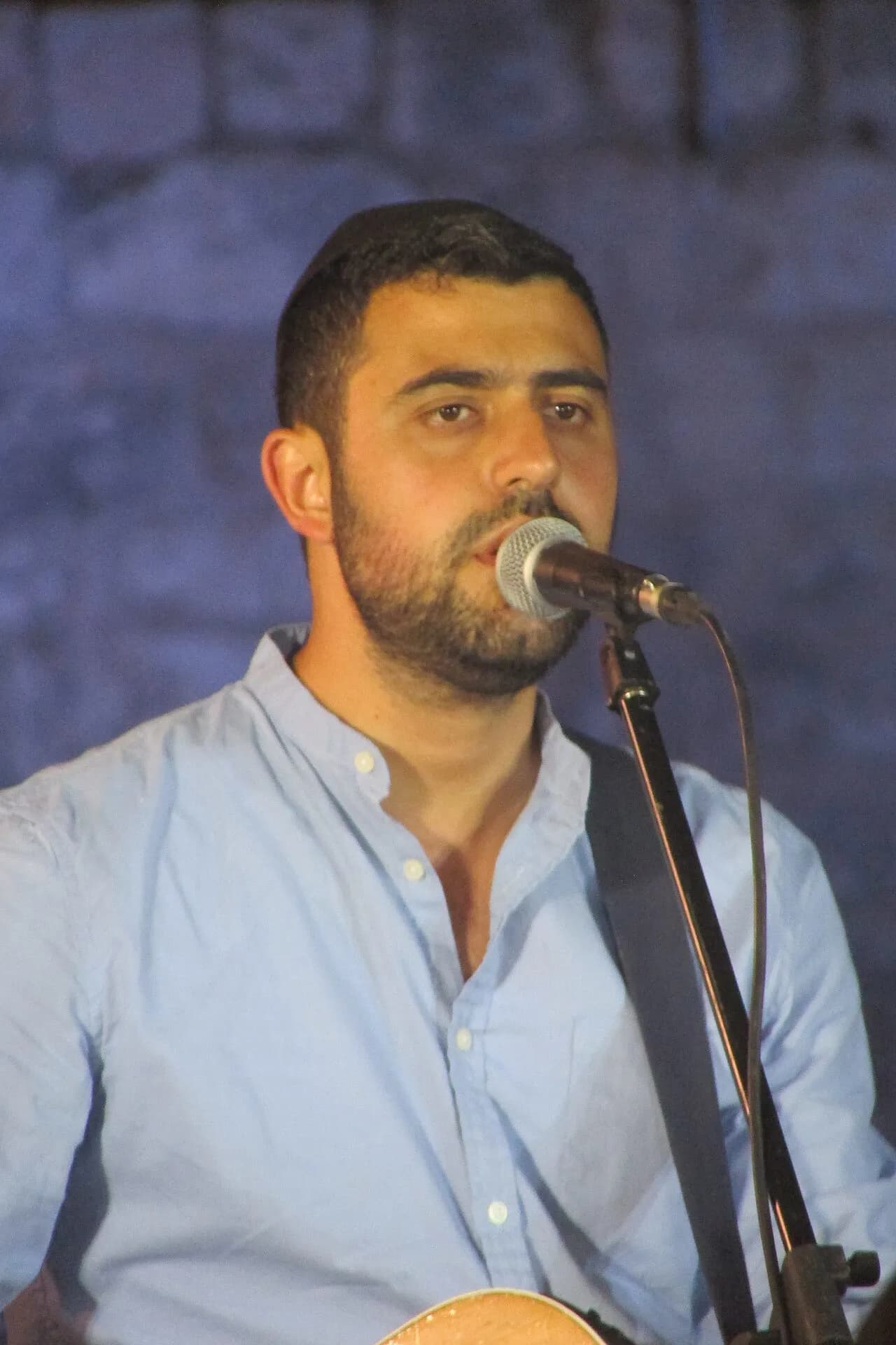 ישי ריבו שר למיקרופון עם גיטרה, בחולצה תכלת, מול קיר אבן, 2017