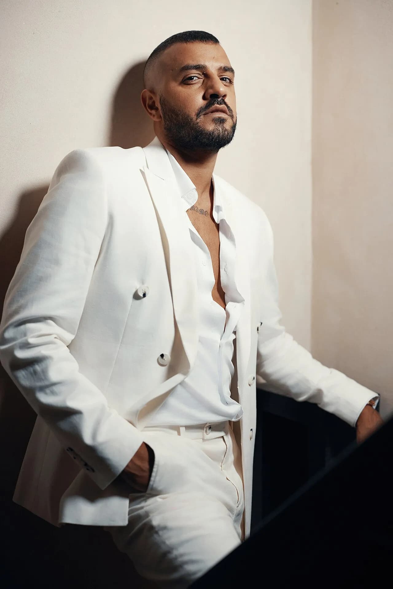 Itay Levi in a white suit and open white shirt, leaning on a wall, 2023