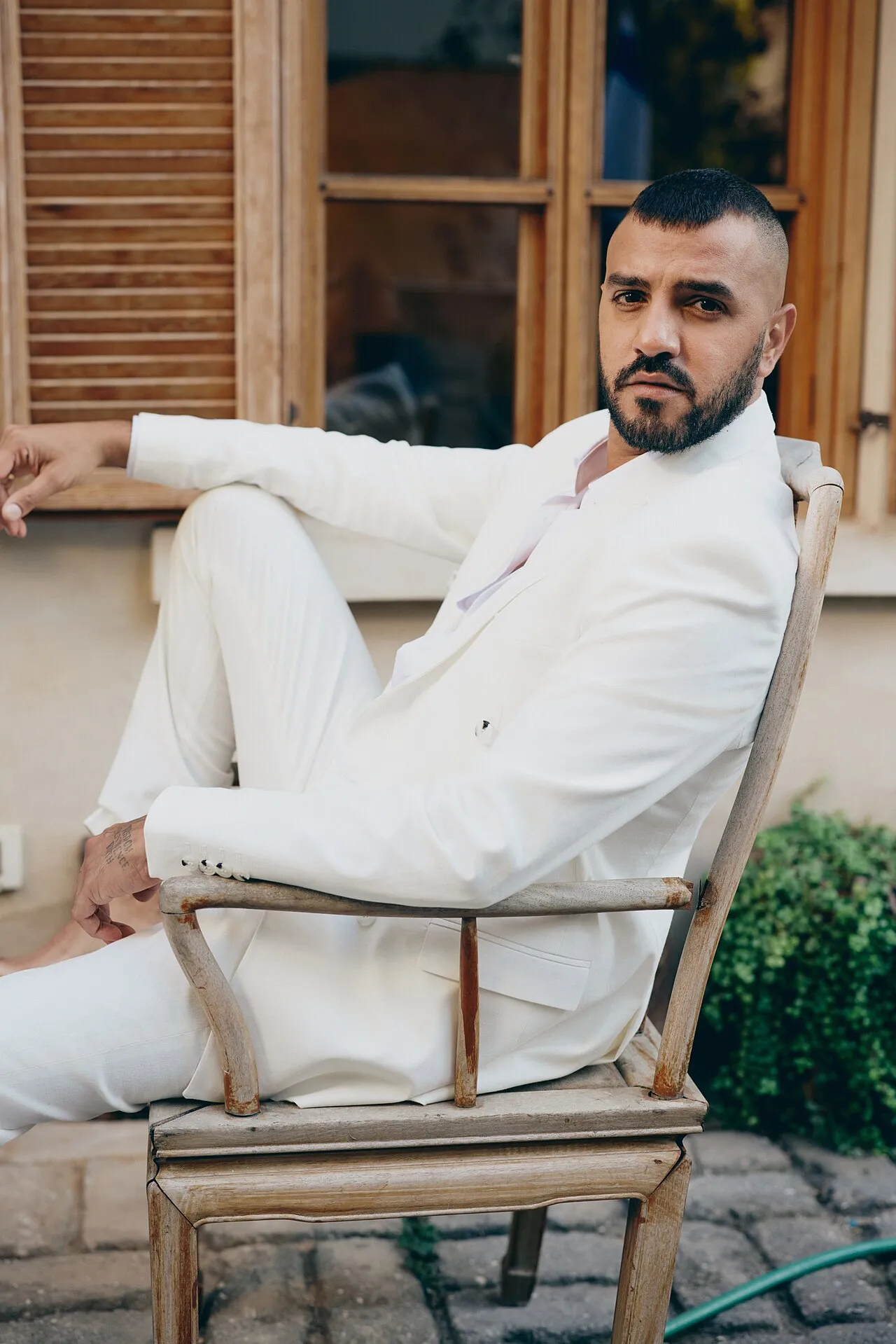 Itay Levi in a white suit, sitting on a wooden chair outside a house with brown shutters, 2023