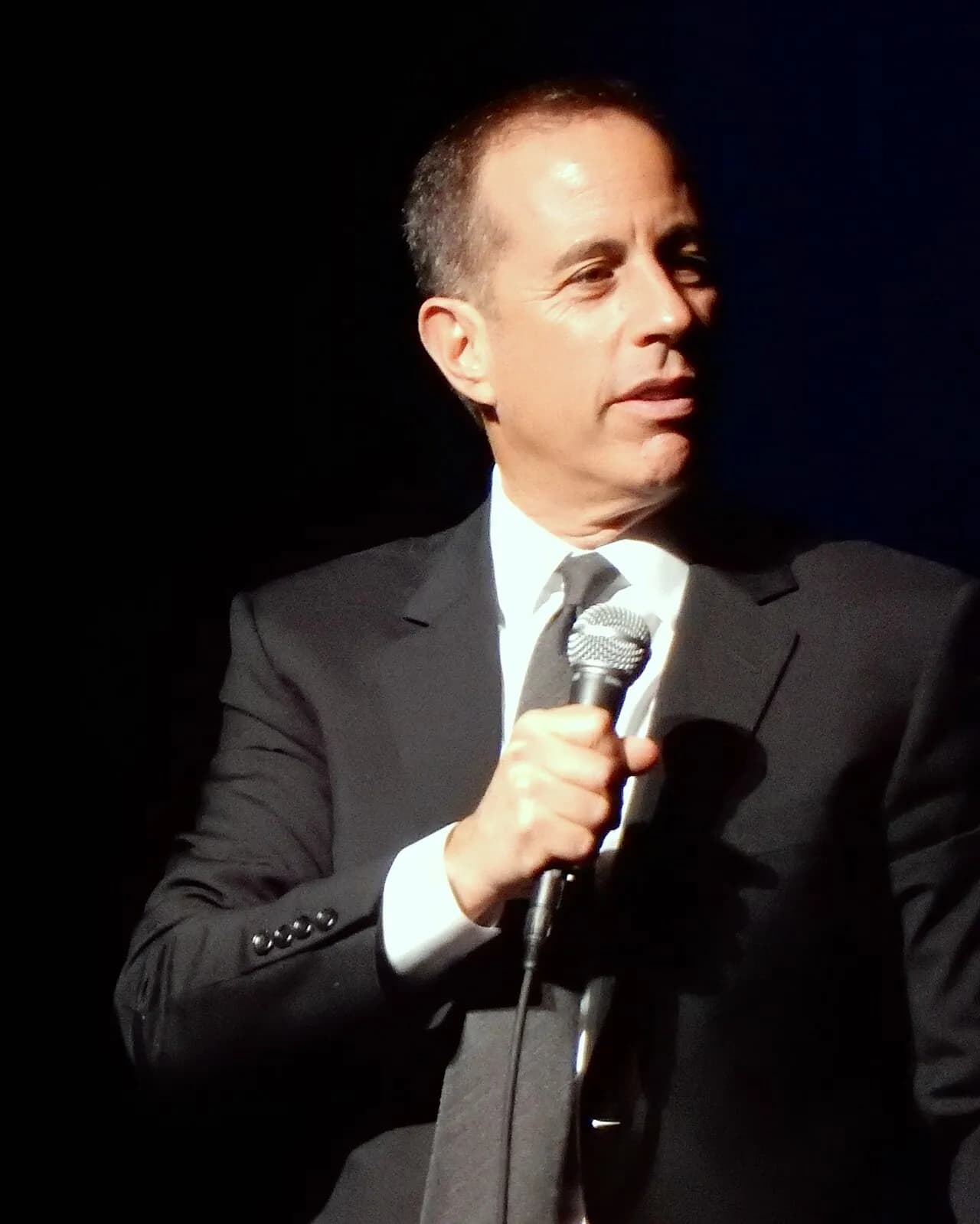 ג'רי סיינפלד על הבמה עם מיקרופון ביד, בחליפה כהה ועניבה, 2016