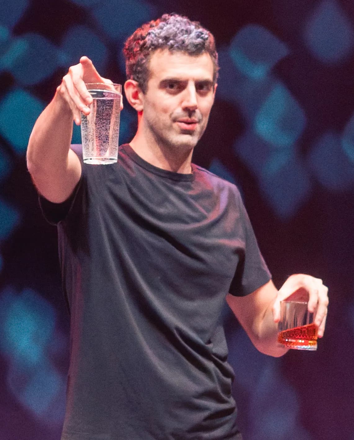 סם מוריל על הבמה בחולצה שחורה, מרים כוס לעבר הקהל, 2025