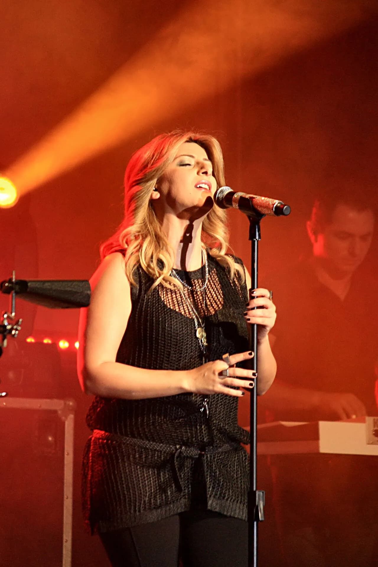Sarit Hadad singing at a microphone stand under orange stage lights, 2013