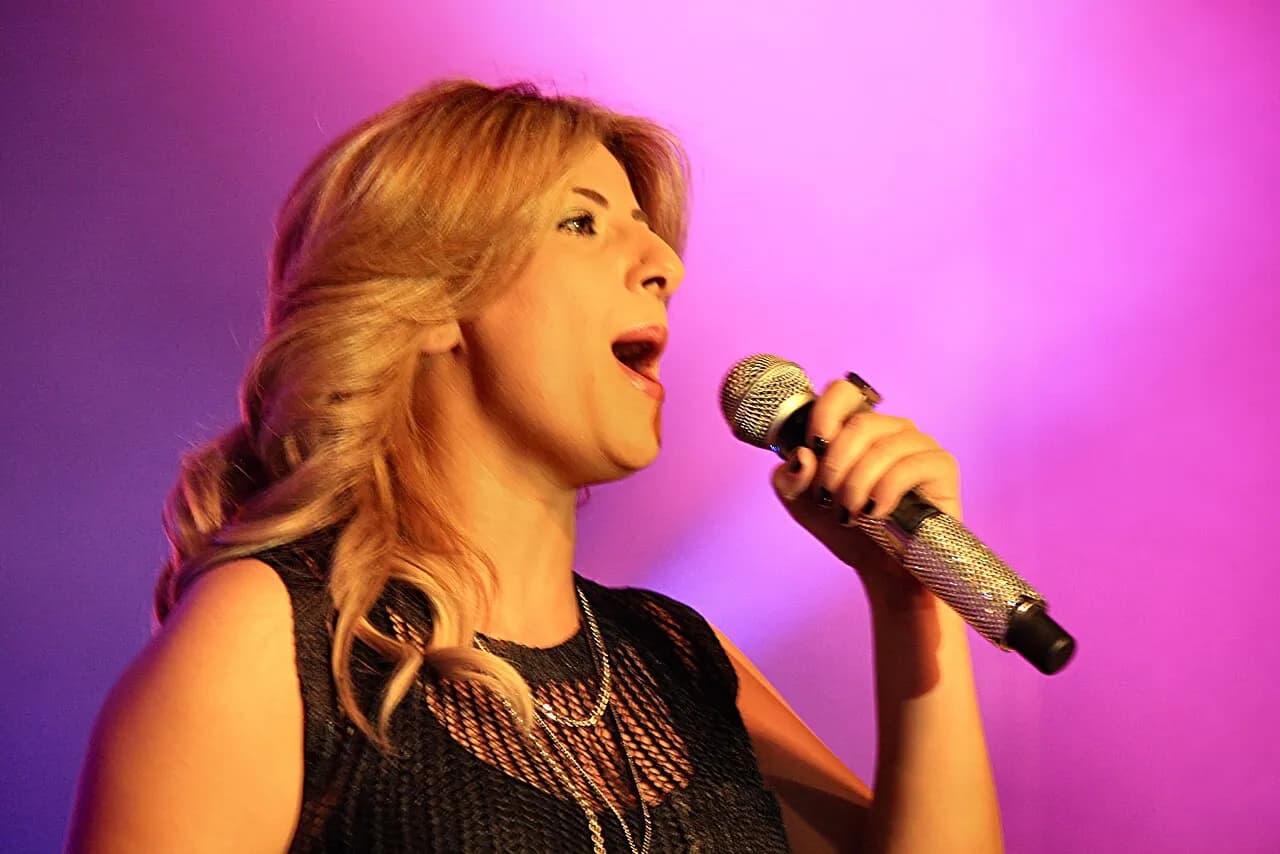 Sarit Hadad singing into a microphone against a pink background, 2013