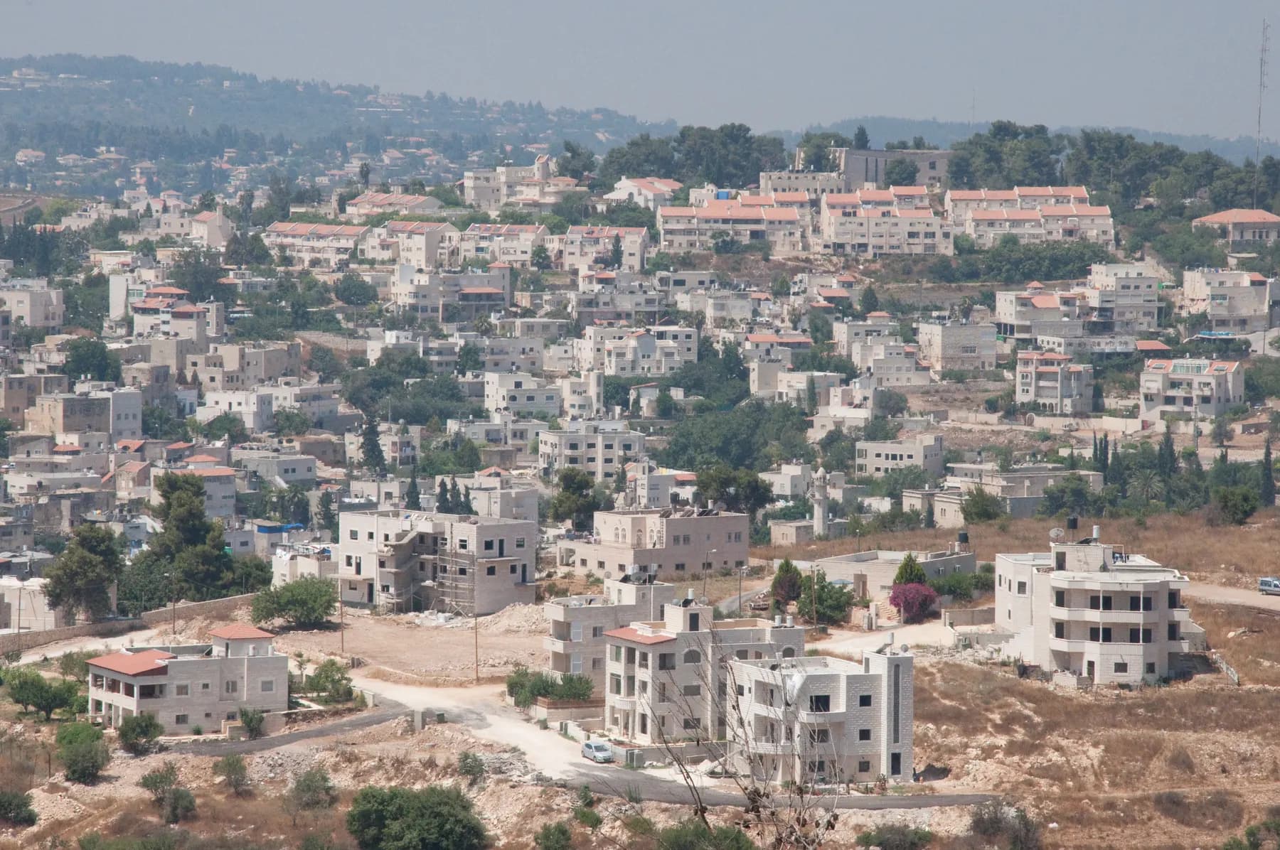 Pale stone houses spread across a hillside in Abu Ghosh, with red-roofed buildings along the ridge above and pine trees between them.