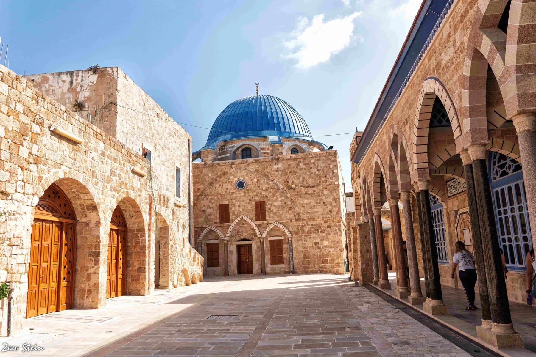 A paved street in Acre's old city, with arched wooden doors, a stone colonnade and a building with a blue dome.