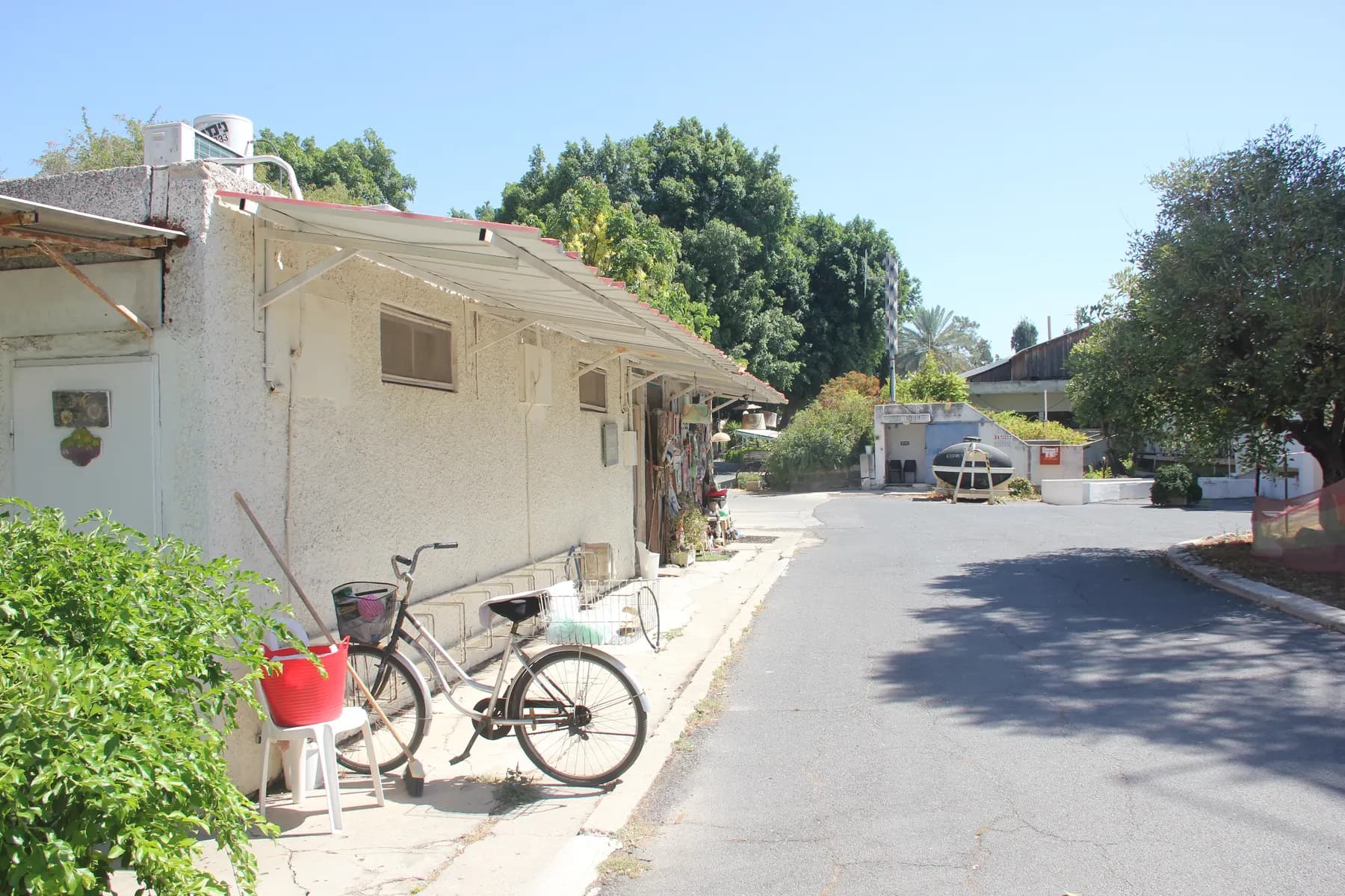 A lane past a long, low white building with bicycles parked by its wall in Afikim, under leafy trees.