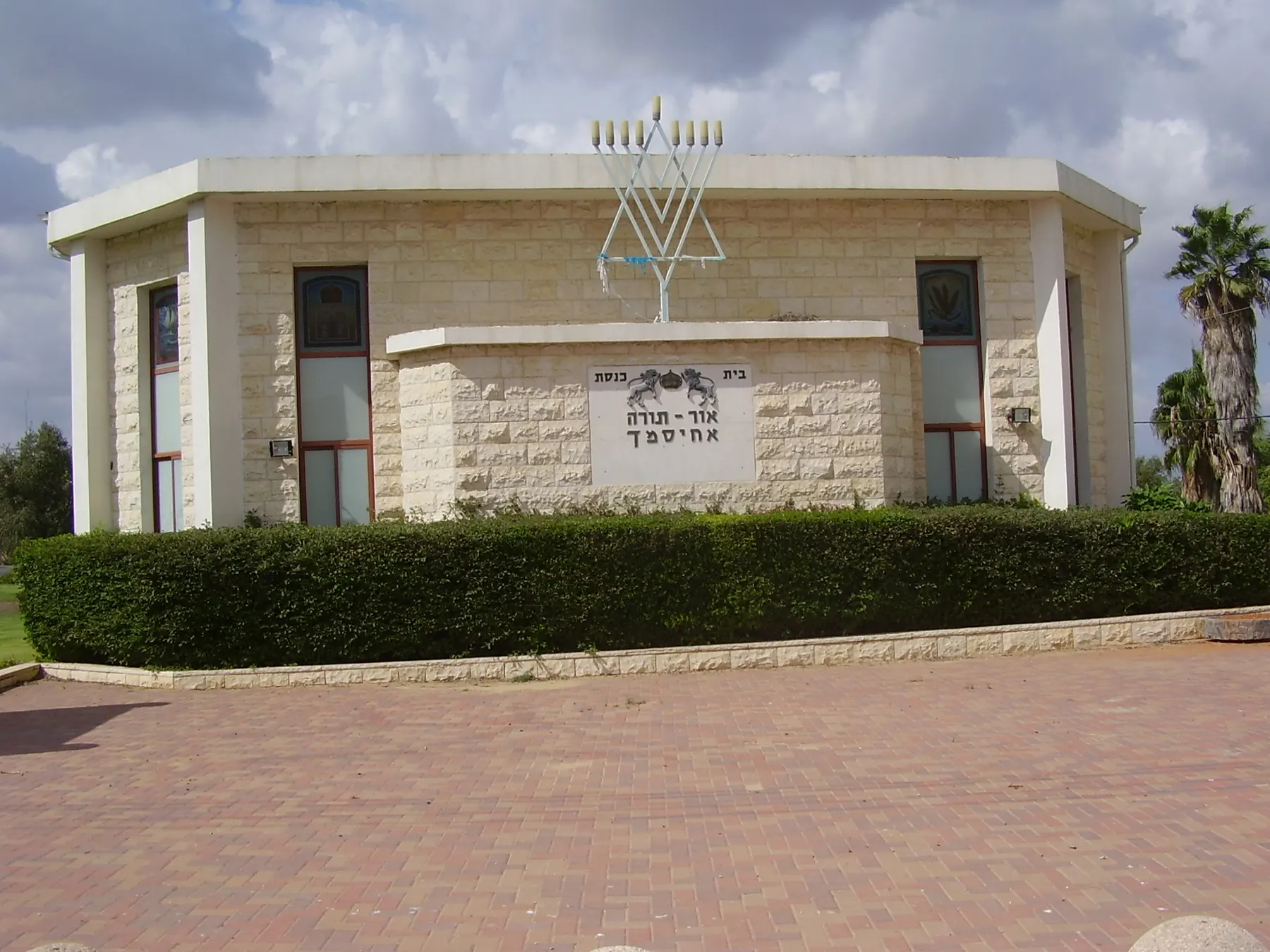 The synagogue of Ahisamakh, a pale stone building with a menorah on its roof, behind a trimmed hedge and a paved square.