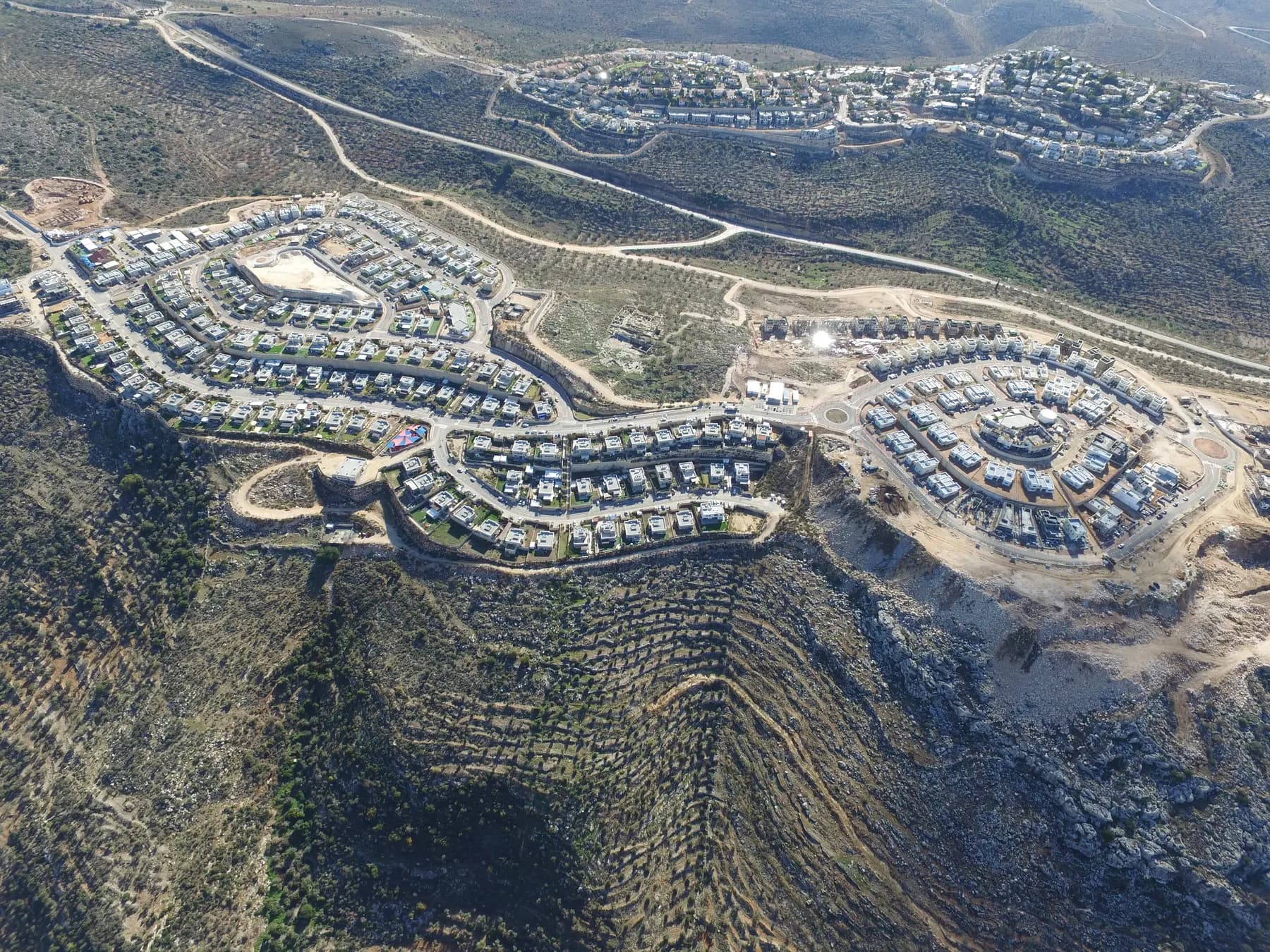 An aerial view of the Leshem neighbourhood of Alei Zahav, rows of new houses along curving streets on a hilltop above terraced slopes, with more houses on the next hill.