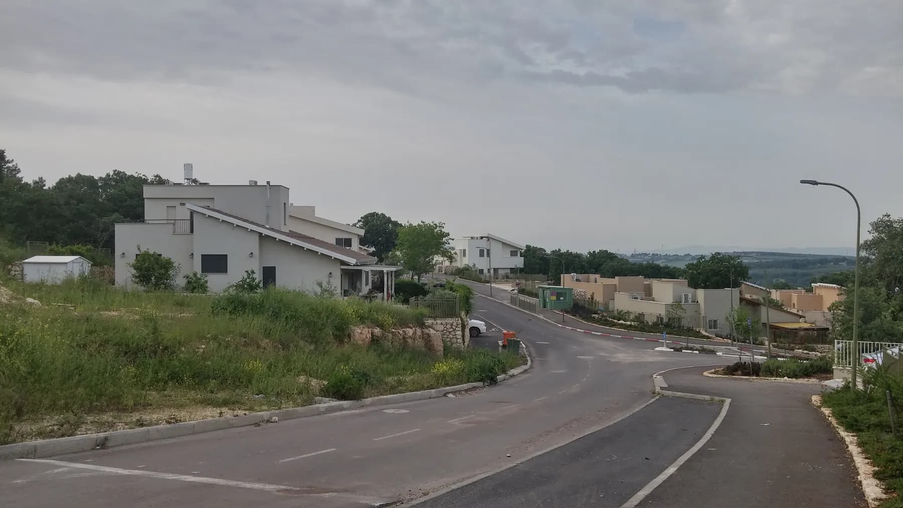 A curving street in Alon HaGalil, with modern white houses on a grassy slope, more houses further down and hills on the horizon under an overcast sky.