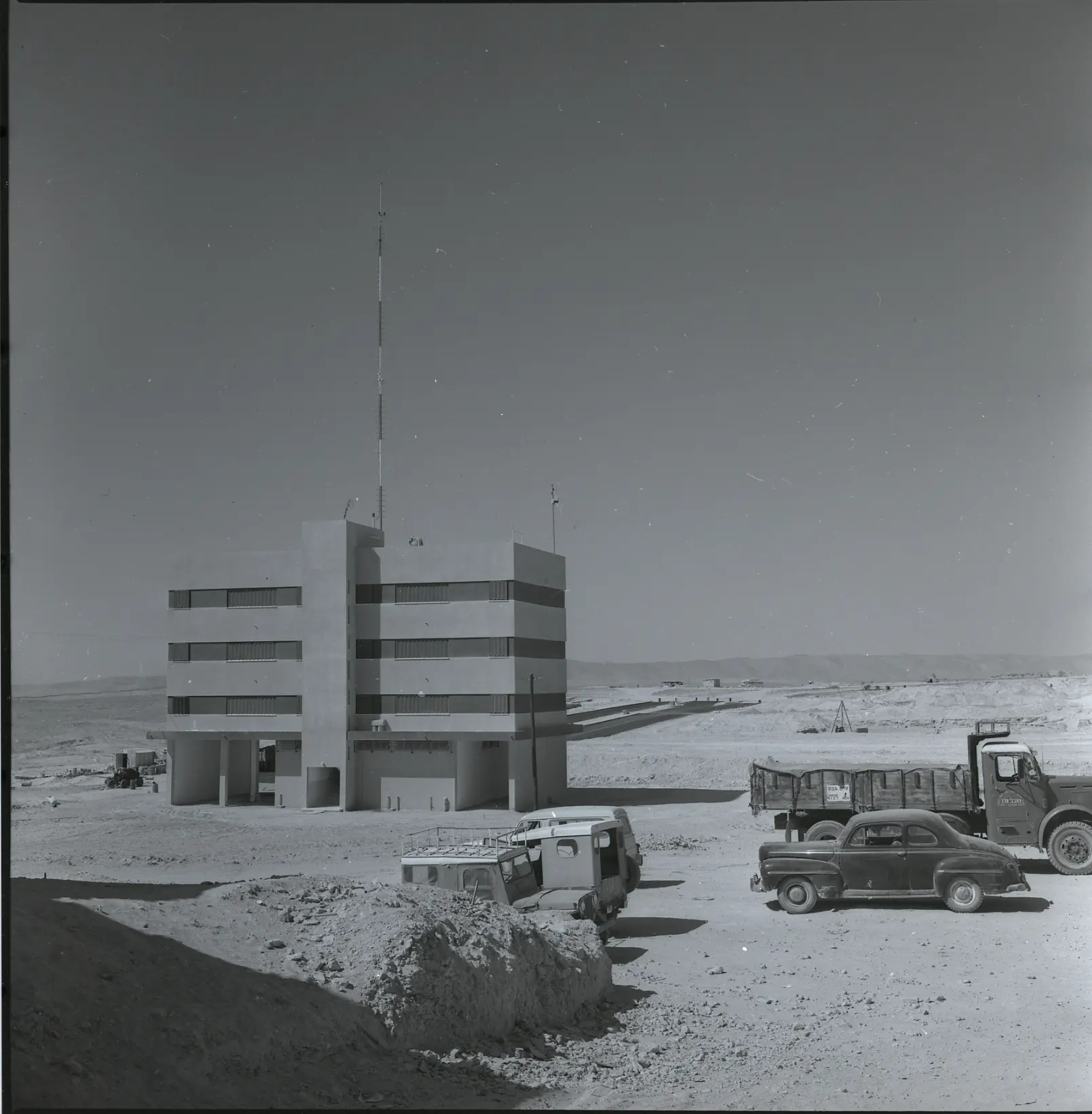An old black-and-white photograph of a new four-storey building on open desert ground in Arad, with old cars and a truck beside it.