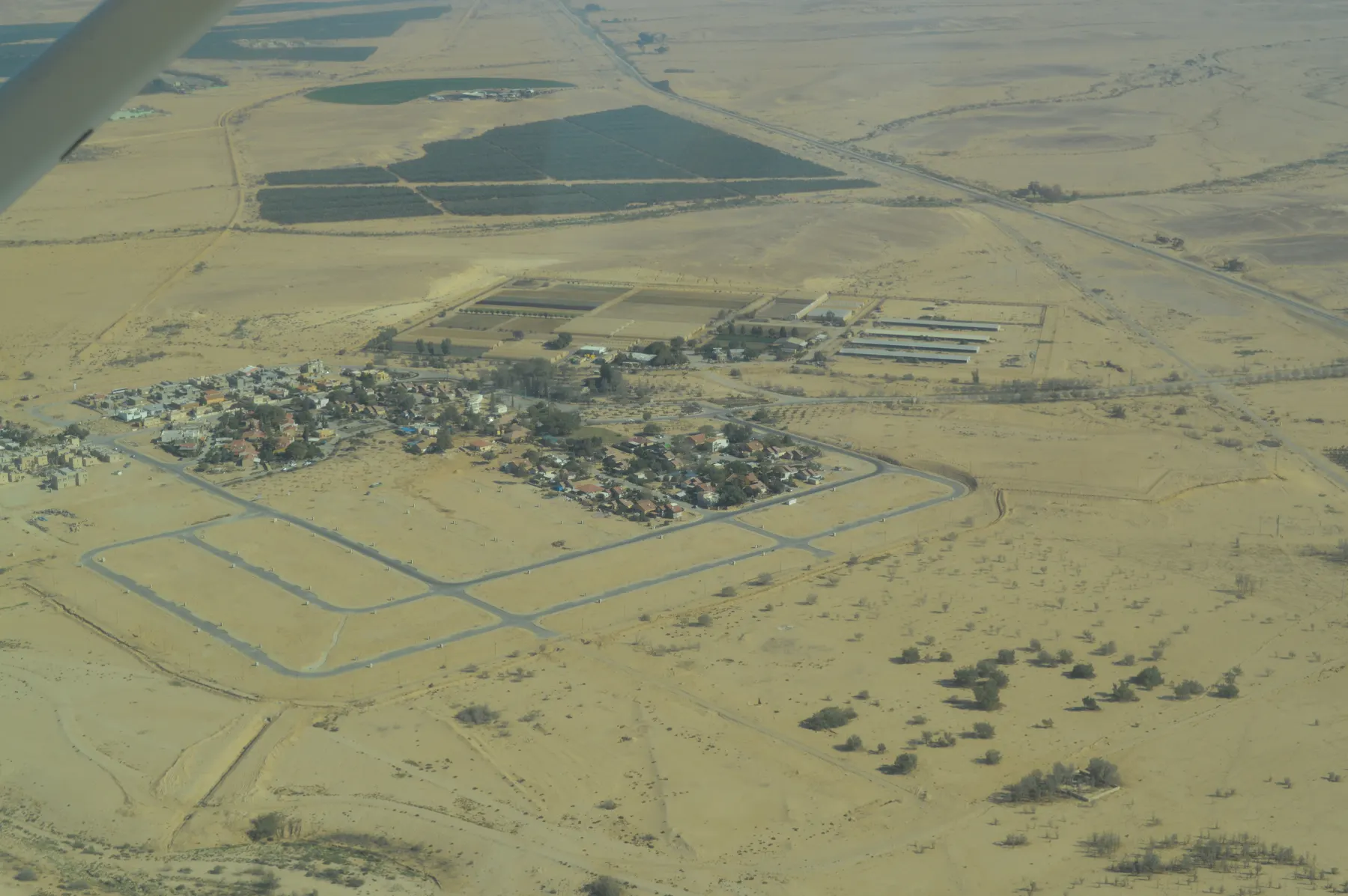 An aerial view of the houses of Ashalim in the desert, with empty streets laid out for new homes in front and farm plots and large dark fields beyond.