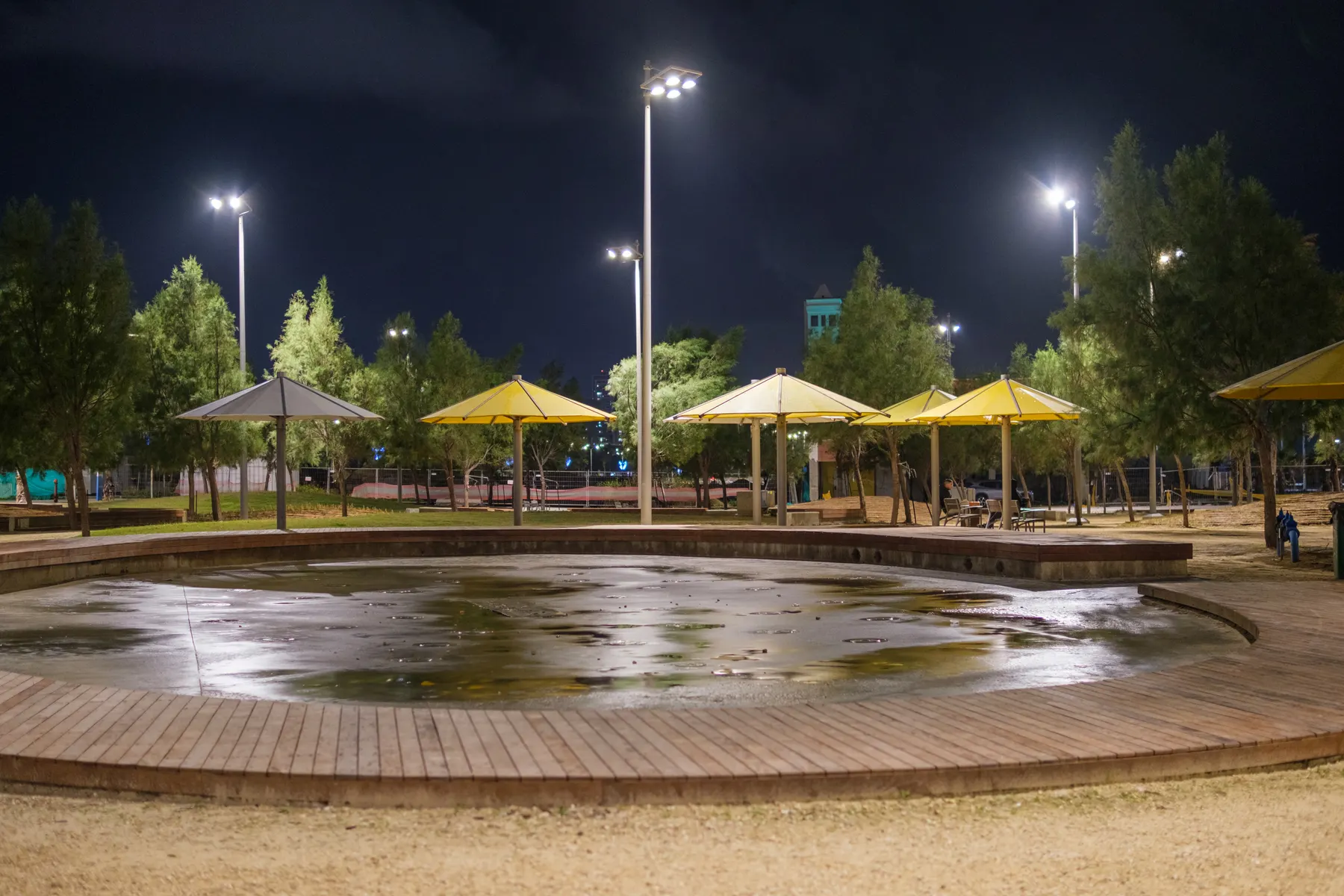 The promenade of Ashdod's Lido and Oranim beaches at night, with lit sunshades and trees around a paddling pool