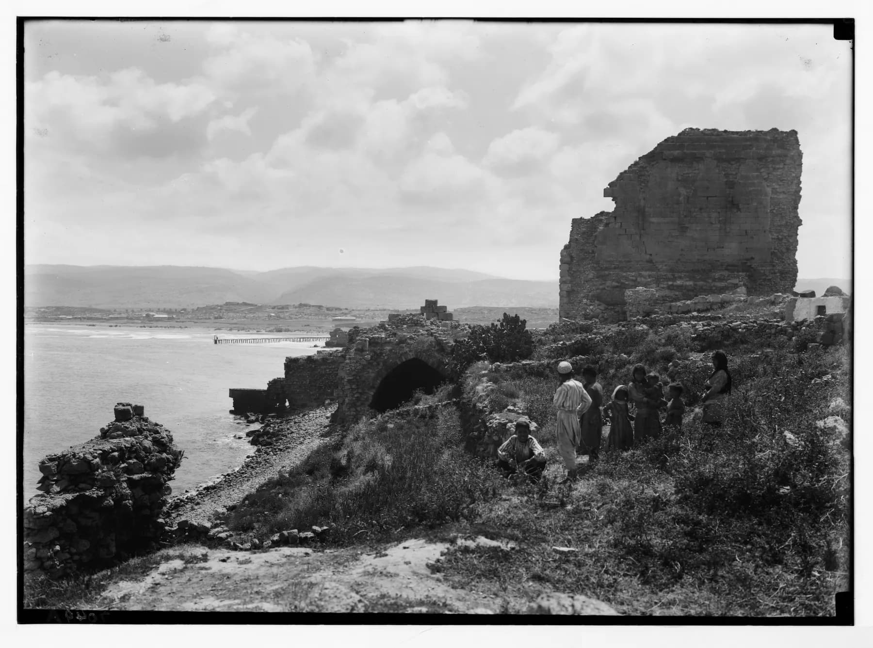 An old black-and-white photograph of a ruined stone tower and arched walls on the shore at Atlit, with a group of people on the grassy slope, a jetty in the bay and hills behind.