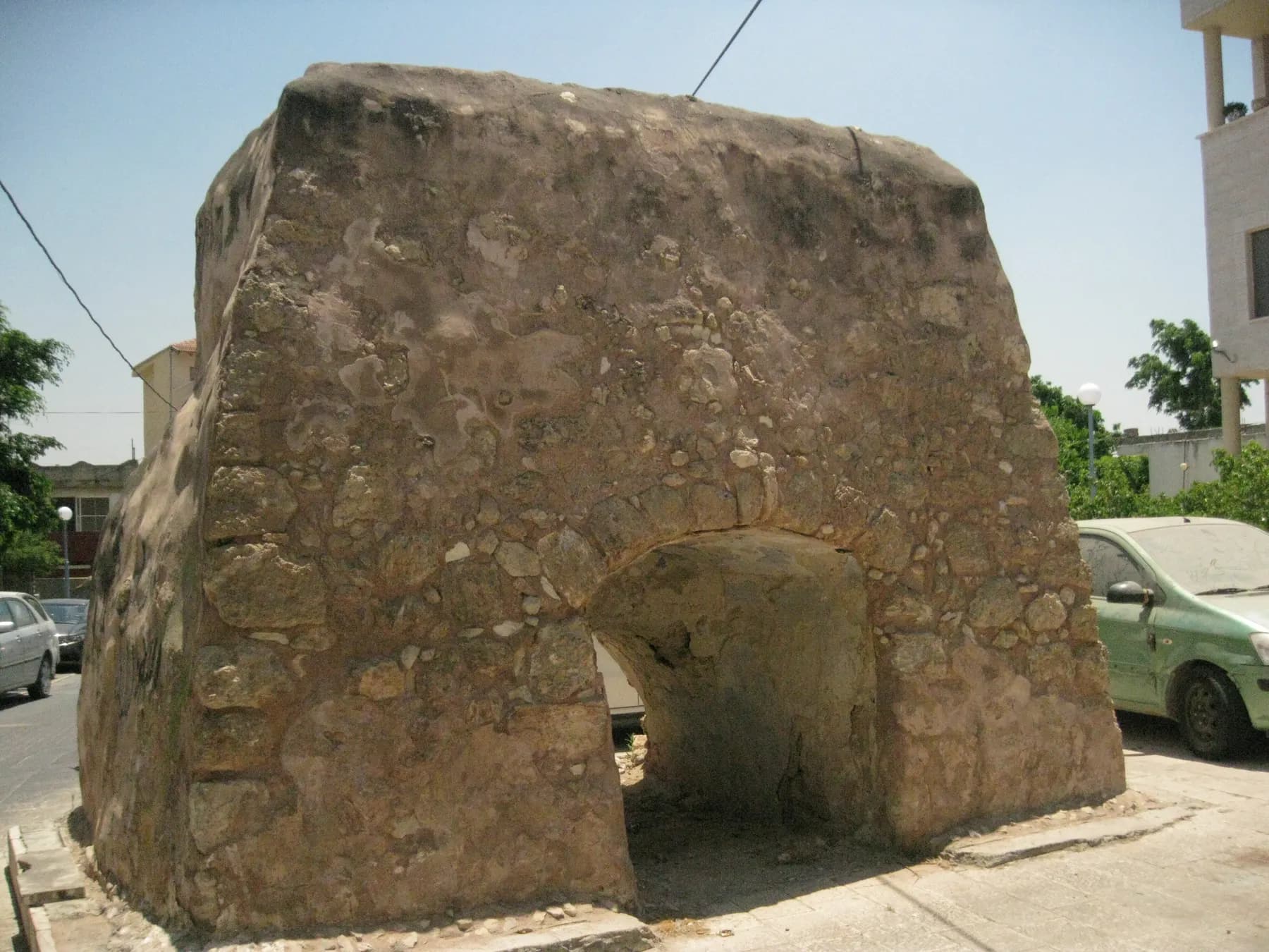 An old vaulted stone structure on a street corner in Baqa al-Gharbiyye, between parked cars and houses.