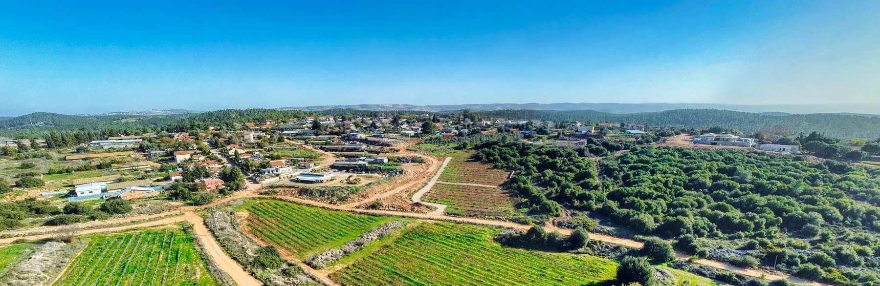 A view from above over Bar Giora, its houses spread along a ridge above terraced green fields, dirt tracks and woodland, with hills on the horizon.
