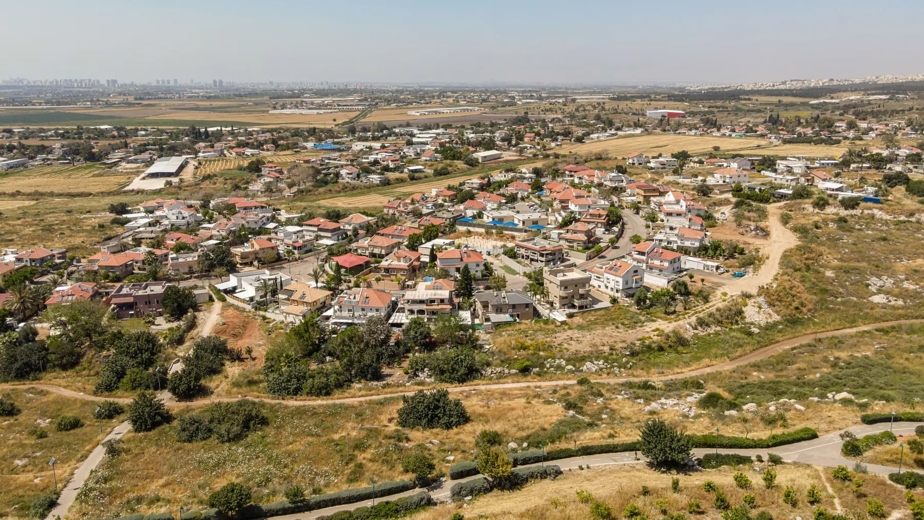 An aerial view over the houses of Bareket, many with red roofs, with dry fields and paths in front and a built-up skyline on the horizon.