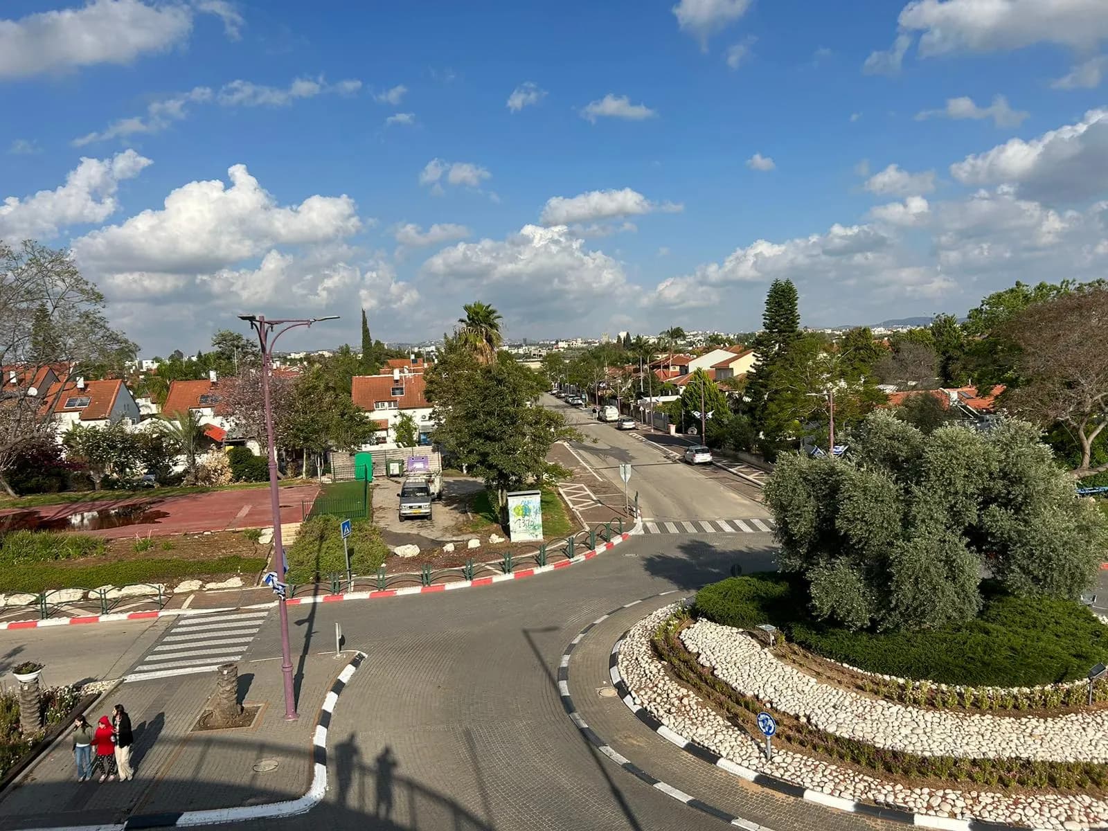 A roundabout planted with an olive tree and white stones in central Bat Hefer, with red-roofed houses, palms and a tree-lined street under a cloudy blue sky.