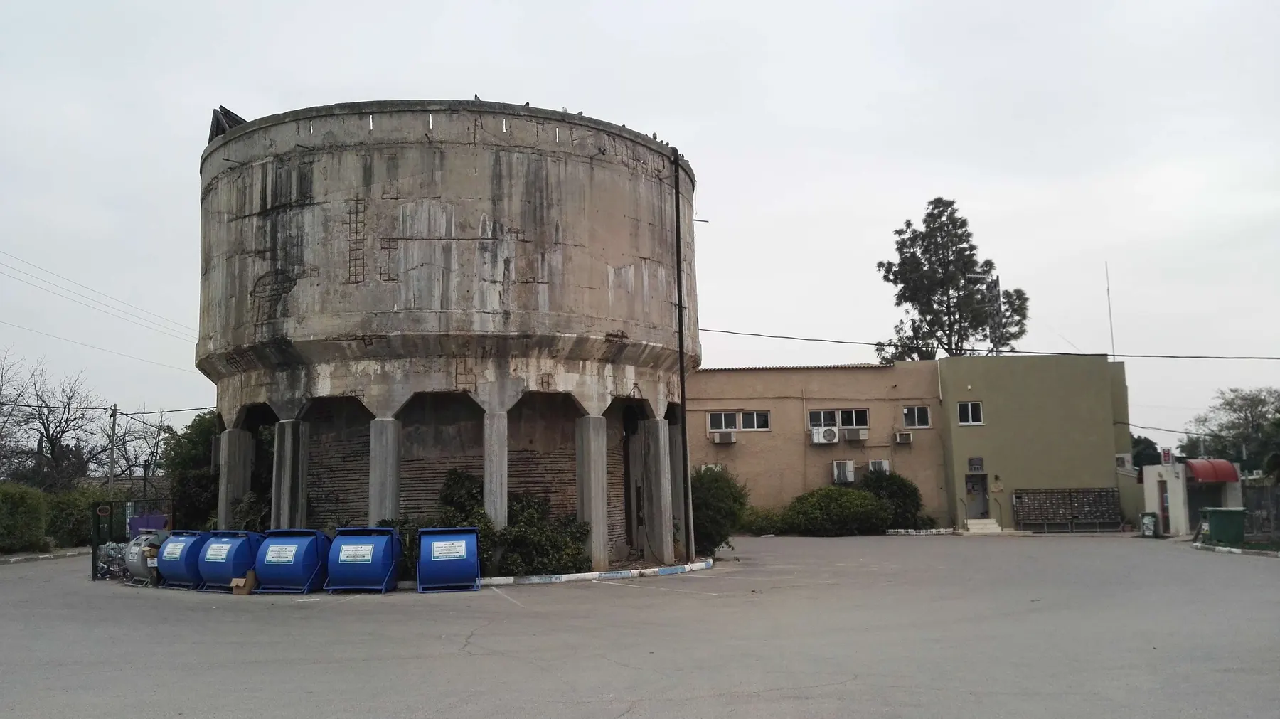 The old water tower of Be'er Tuvia, a wide concrete drum raised on columns, beside a two-storey office building with a row of blue bins in front.