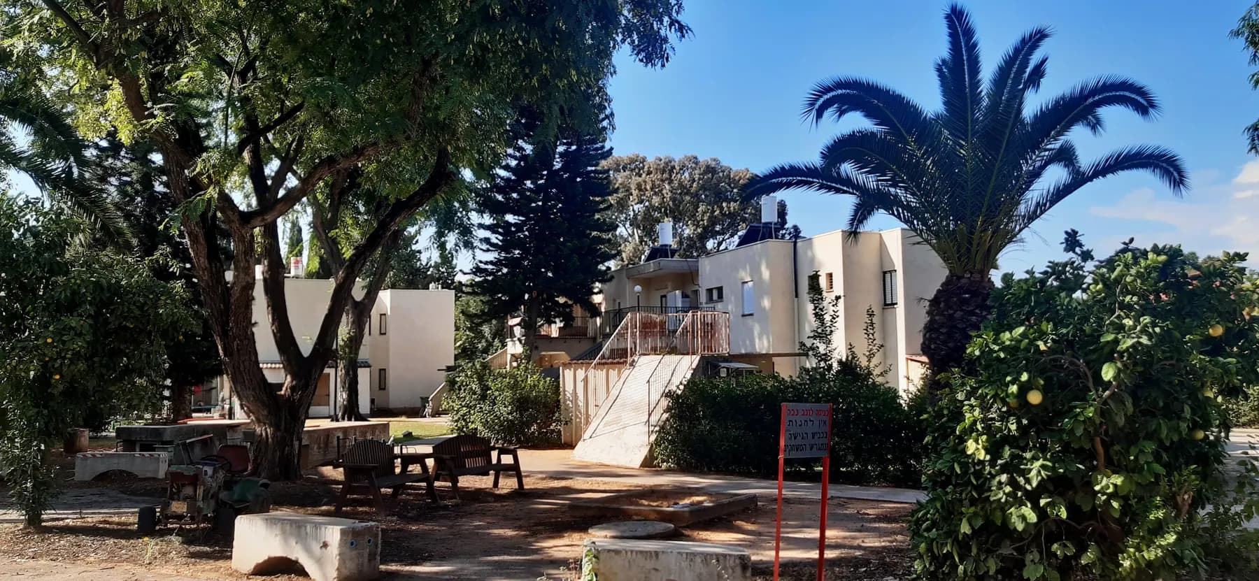 Two-storey houses among trees and a palm in Be'erot Yitzhak, with garden tables and benches in a shaded yard.
