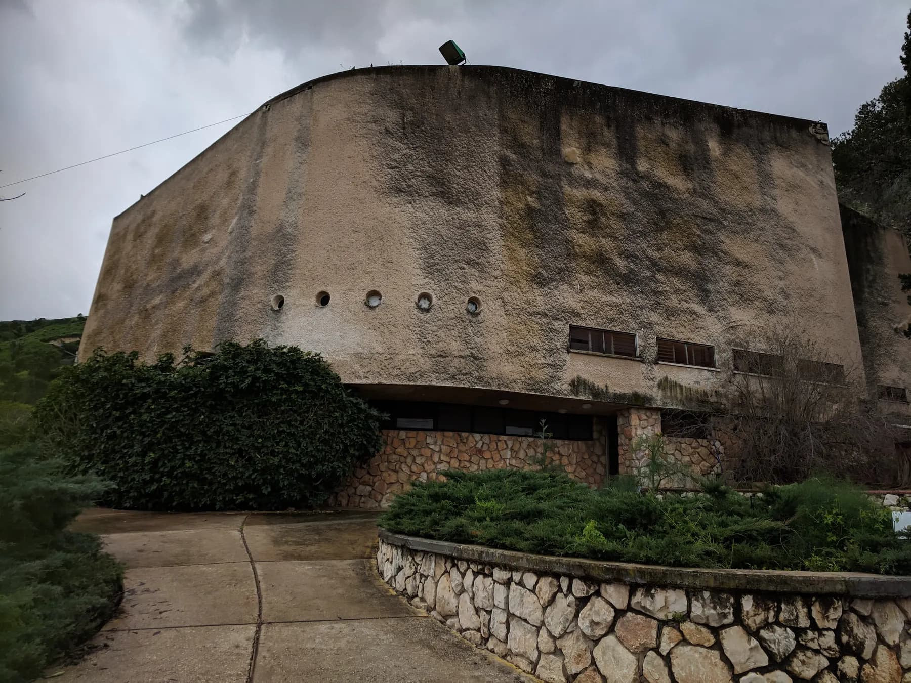 The community hall of Kibbutz Beit Alfa, a large curved concrete building with a row of small round windows, with shrubs and a low stone wall in front.