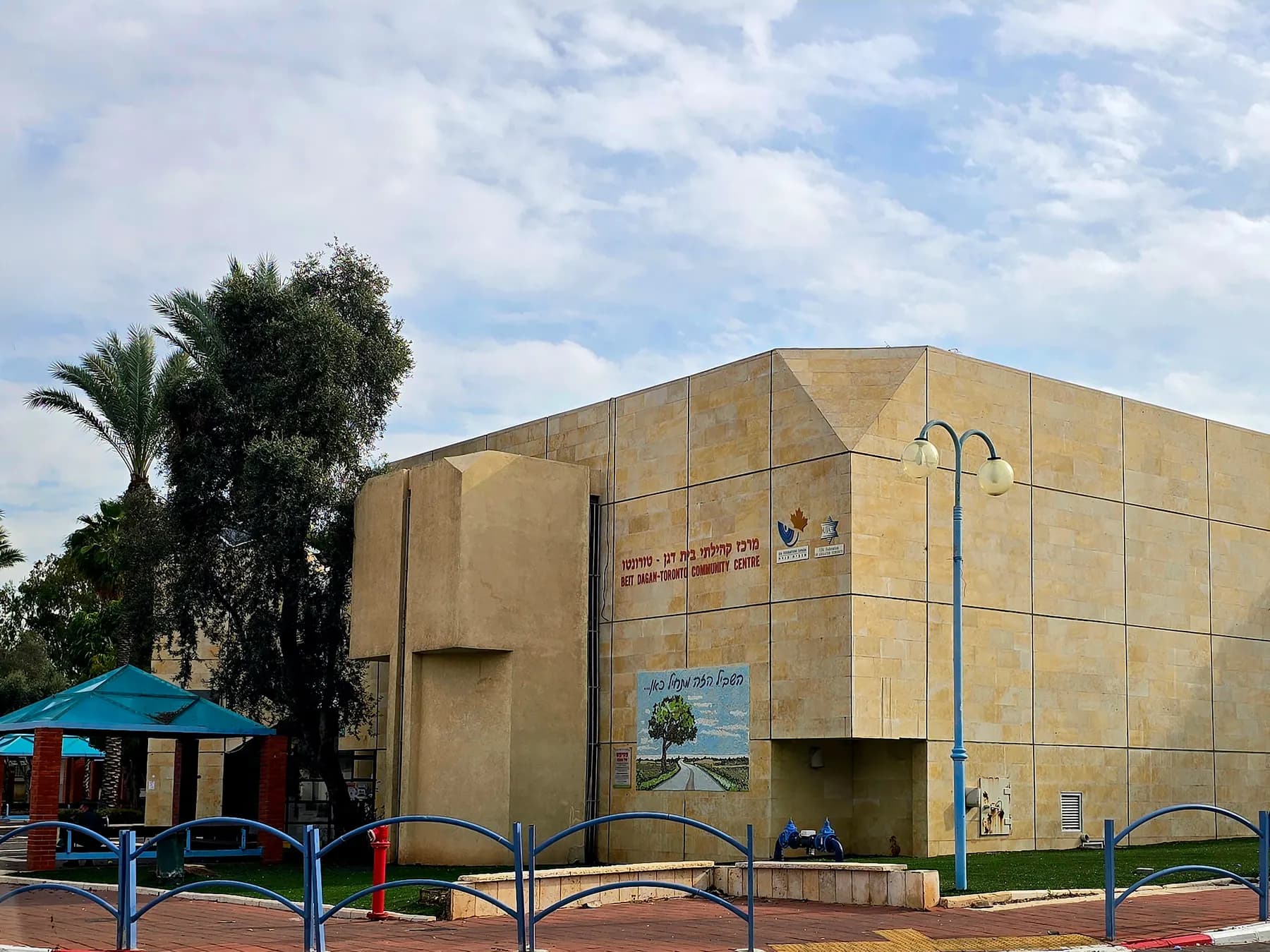 The community centre of Beit Dagan, a square building clad in pale stone, with palm trees, a street lamp and a shaded playground in front.
