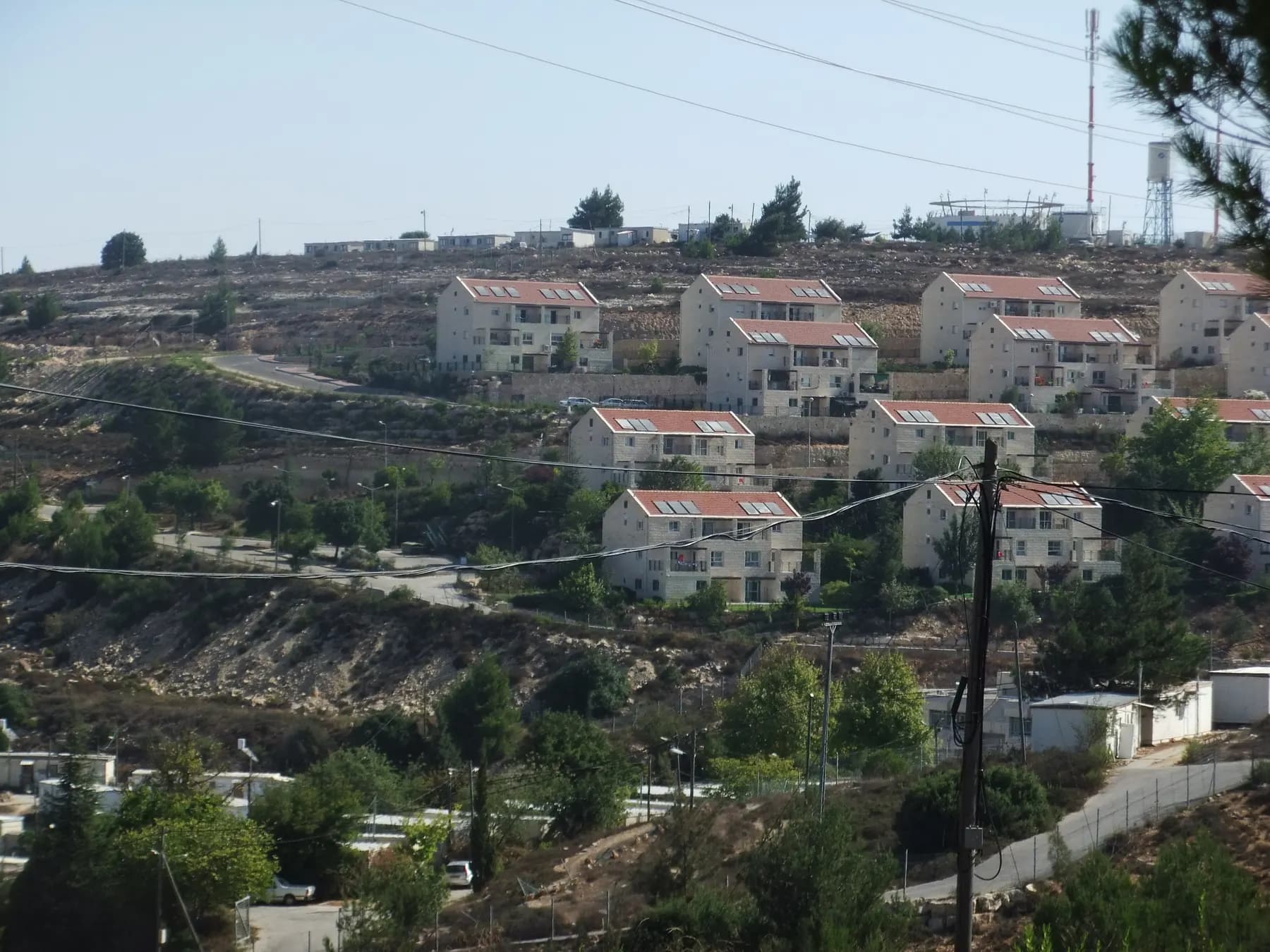 Red-roofed apartment buildings on a terraced hillside in Beit El, with a water tower and an antenna on the hilltop above.