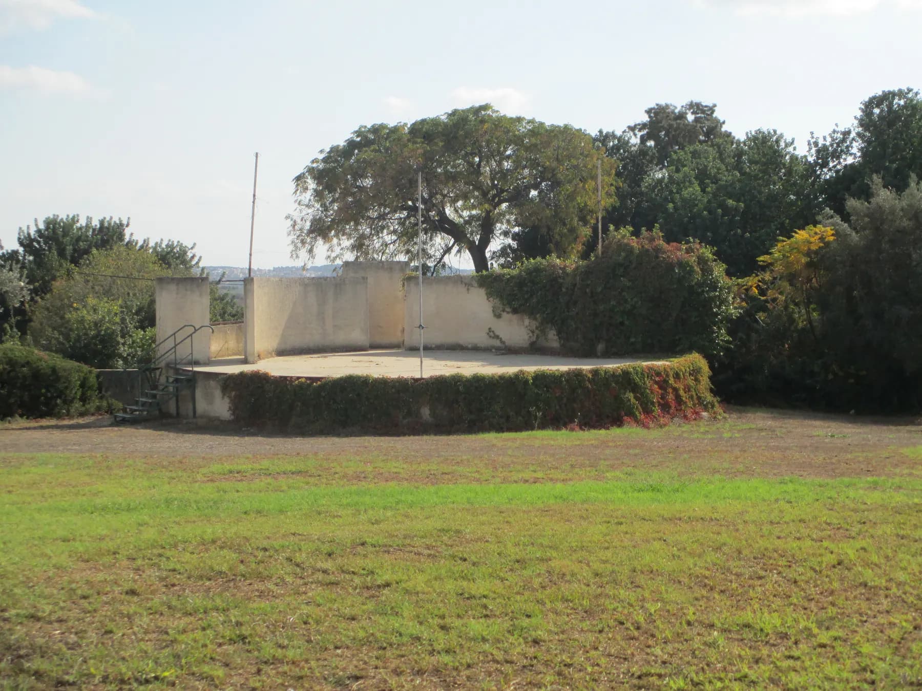 A round open-air stage with low walls at the edge of a lawn in Beit HaEmek, with large trees behind it.