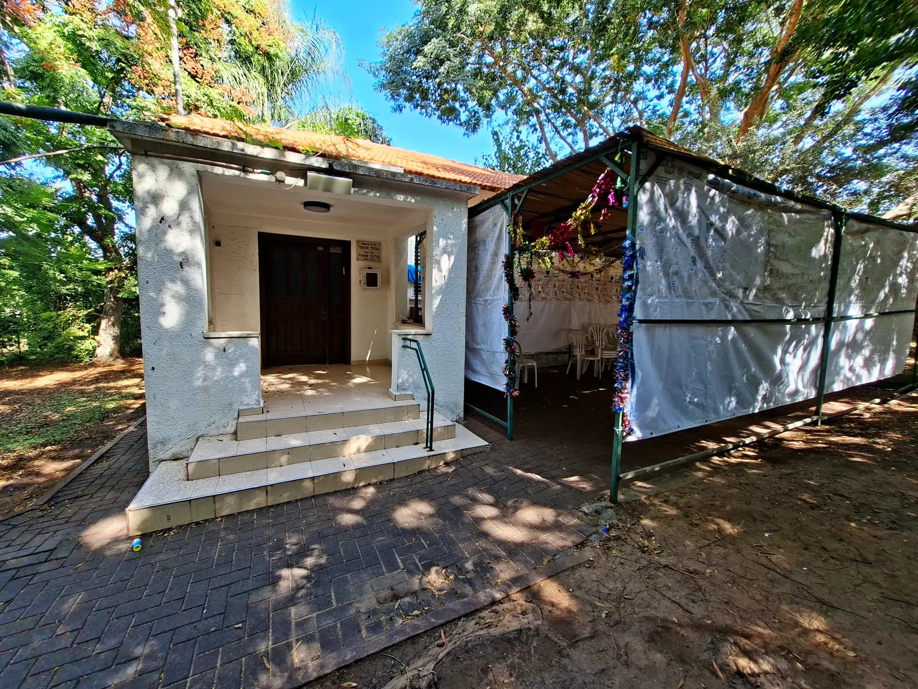 The small white synagogue of Beit HaLevi, with steps up to its doorway and a sukkah with white cloth walls beside it, under tall trees.