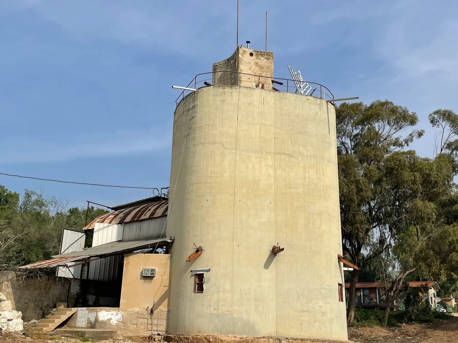 An old round concrete tower with a small square room on top beside a workshop shed in Kibbutz Beit HaShita, with trees around it under a blue sky.