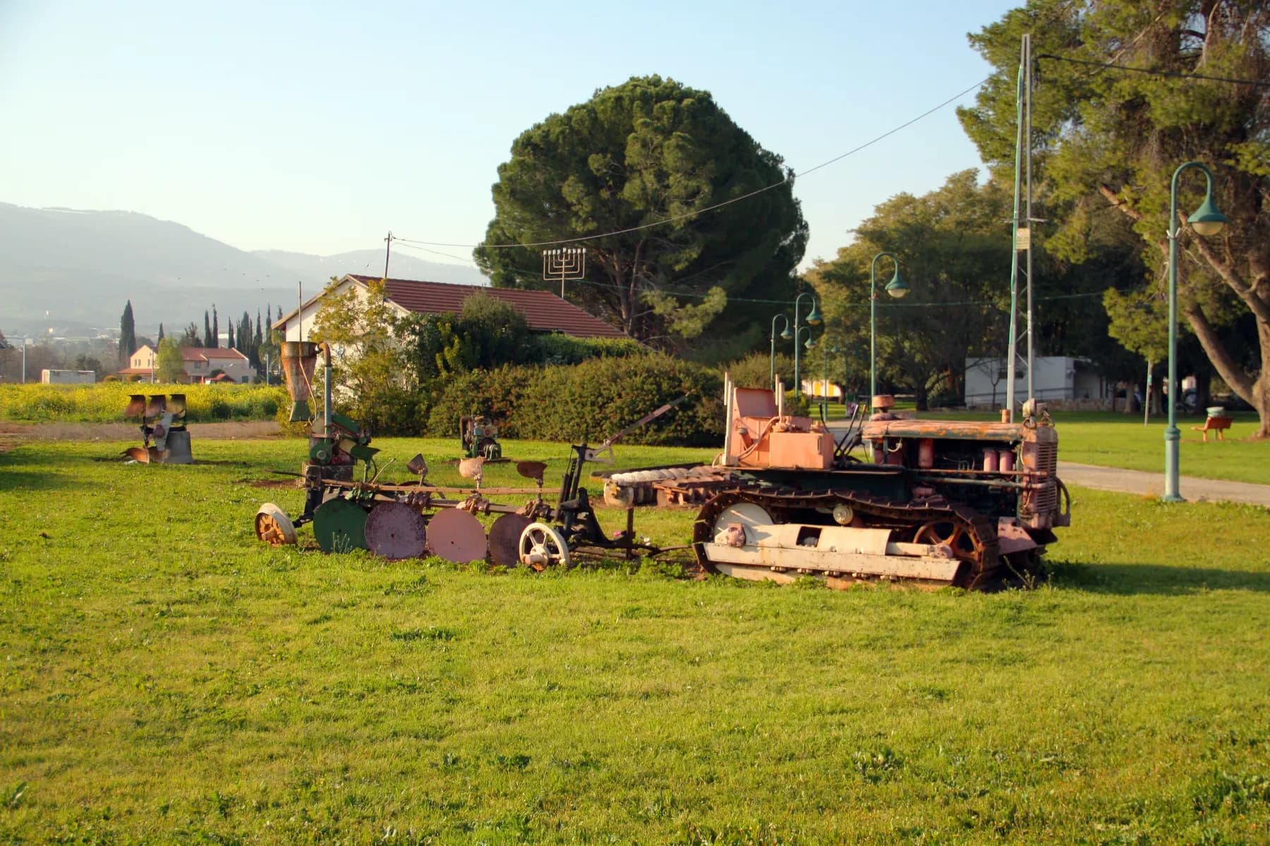 An old rusty crawler tractor and ploughs displayed on a lawn in Beit Hillel, with a red-roofed house, a large pine tree and hills behind.