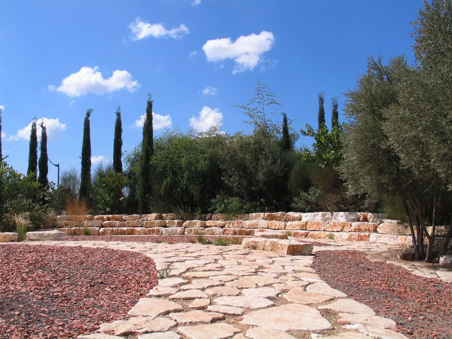 A garden with a stone path, low stone walls, cypresses and olive trees under a blue sky in Beit Nehemia.