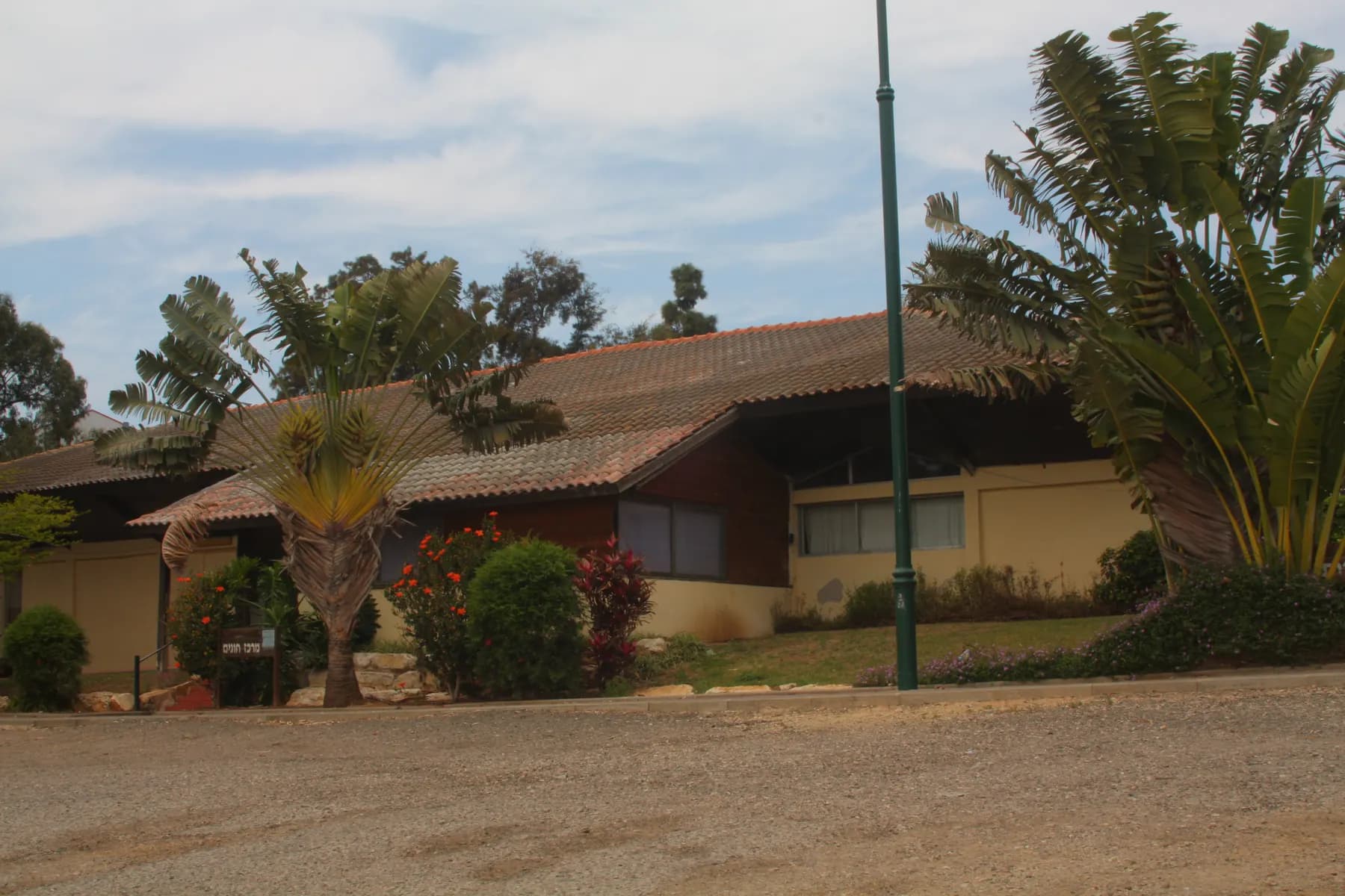 A low building with a tiled roof among palms and flowering shrubs in Beit Yehoshua, across a gravel yard.