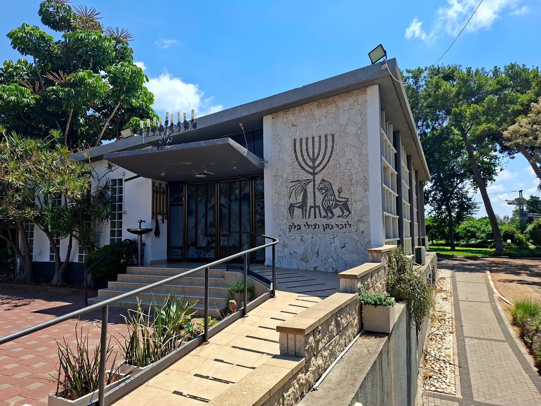 The synagogue of Beit Yitzhak-Sha'ar Hefer, a flat-roofed building with a large mosaic of a menorah and two lions beside its glass entrance, reached by steps and a ramp.