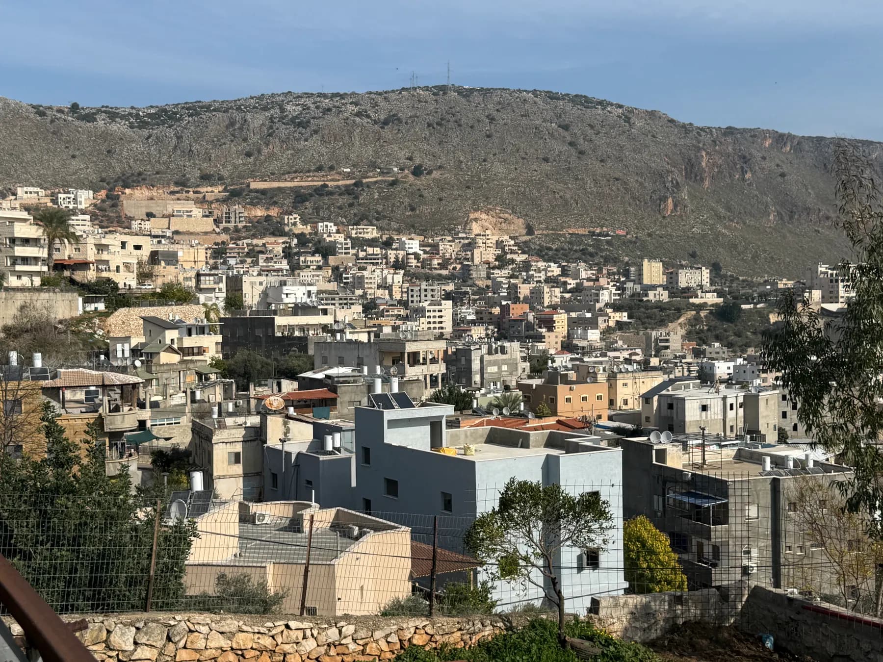 Houses spread up the slope below a steep rocky ridge in Bi'ina, seen over newer buildings in the foreground.