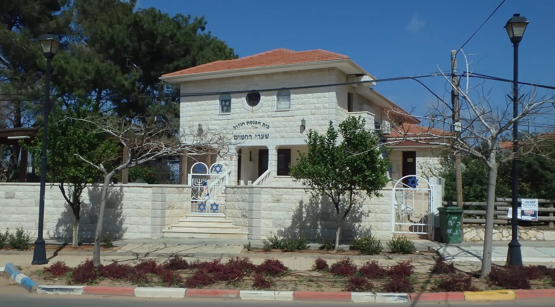 A white stone synagogue with a red tiled roof and a garden in front, on a street in Binyamina-Givat Ada.