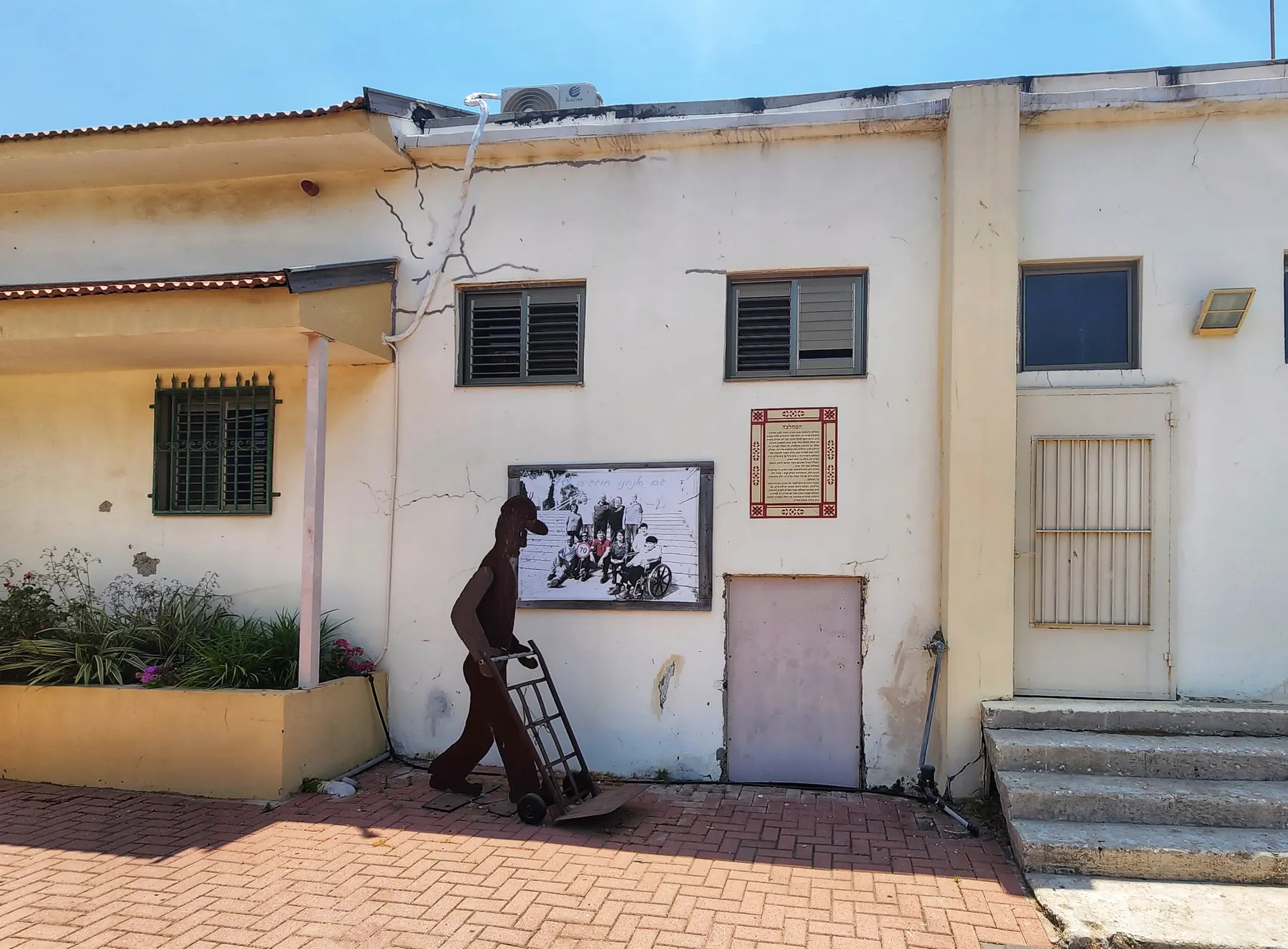 מבנה ישן בצבע שמנת בביצרון עם דמות מתכת חלודה של אדם הדוחף עגלת משא, תמונת קיר גדולה וערוגת פרחים לפניו.