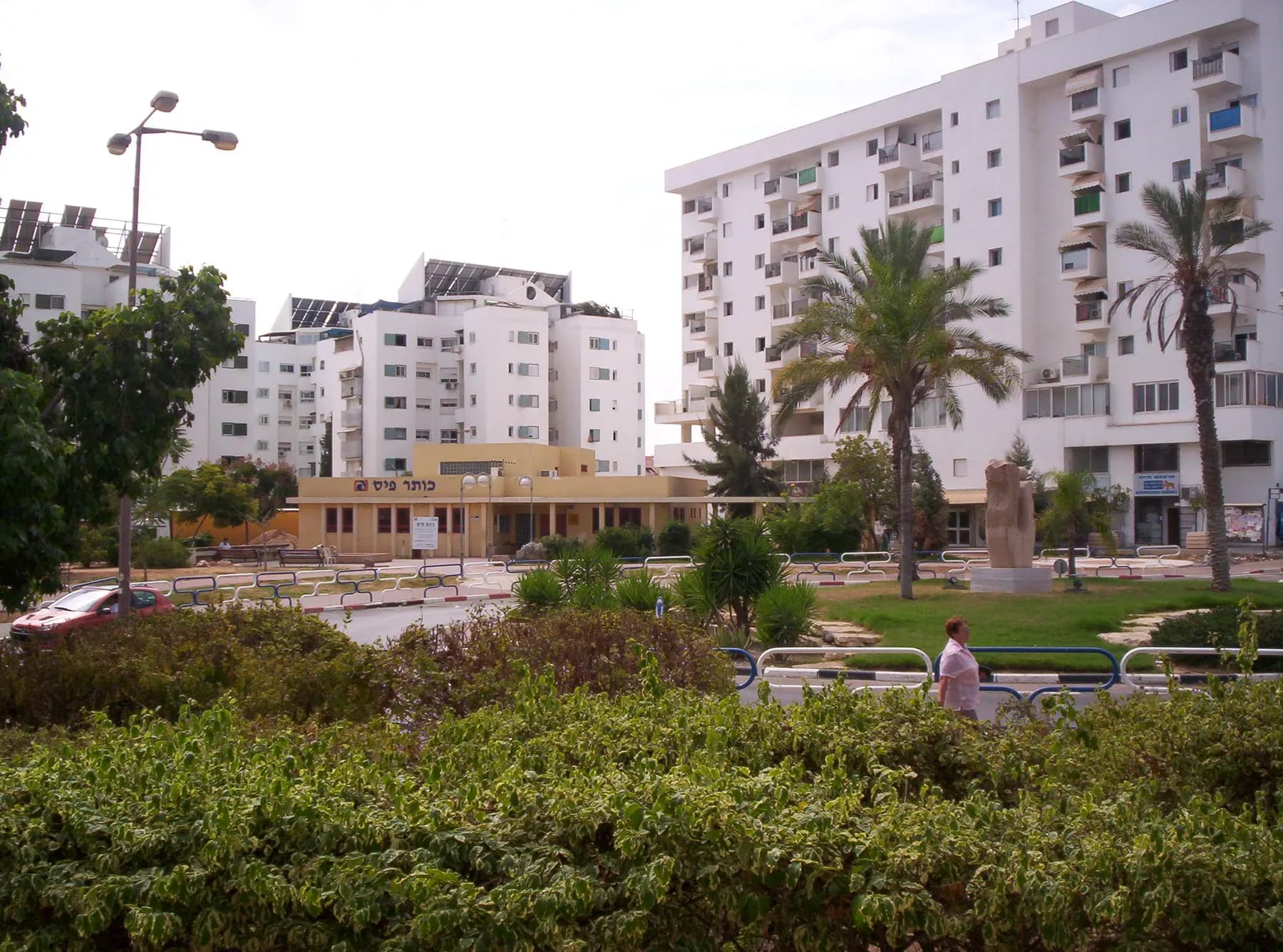 A low shopping centre beside a garden with palm trees in Bnei Ayish, with tall white apartment blocks behind it.
