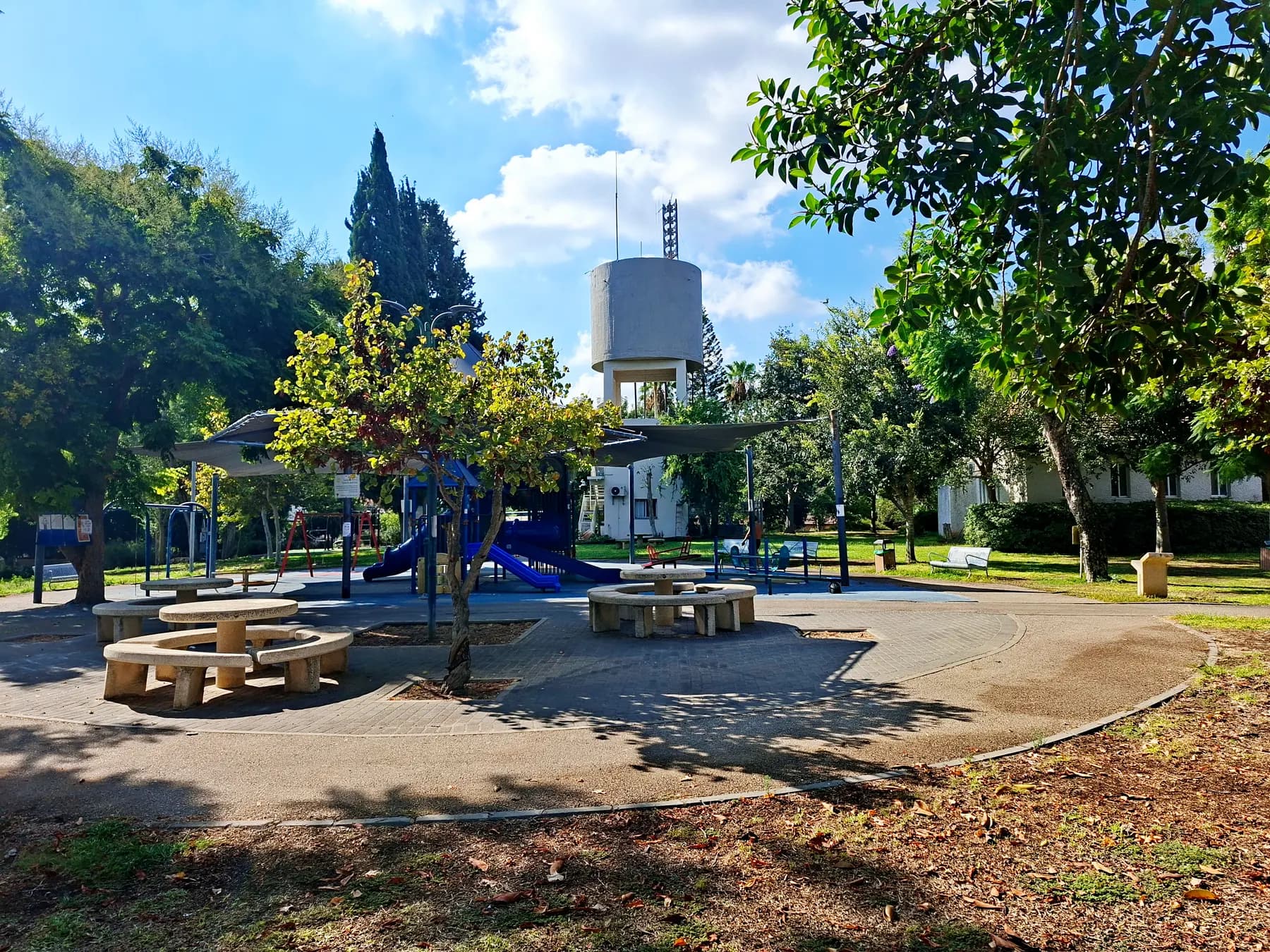 A playground with picnic tables under shade sails in a park in Bnei Dror, with the village water tower rising behind the trees.