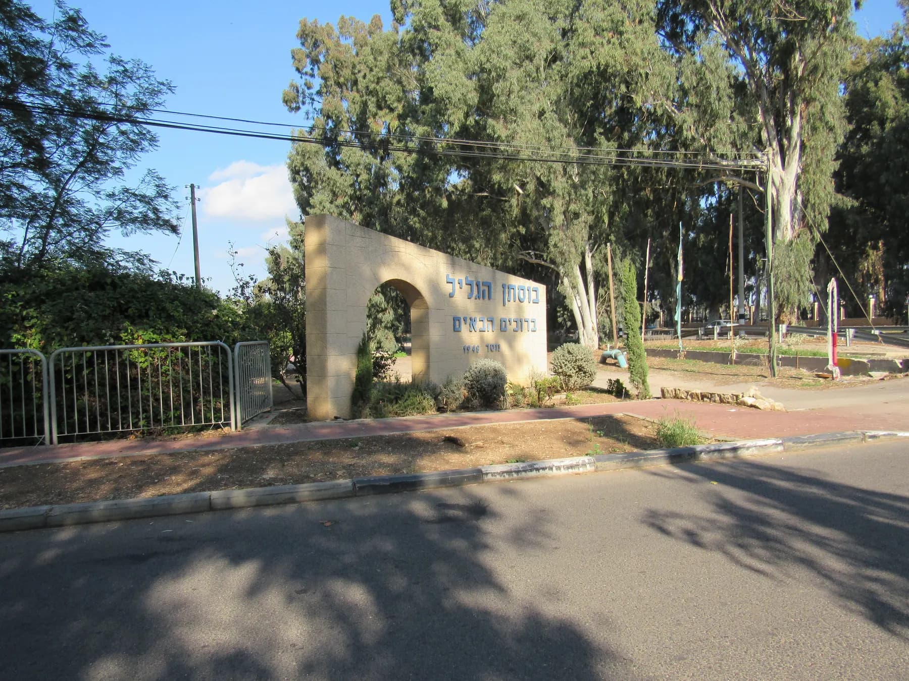 The entrance to Bustan HaGalil, a pale stone wall with an arch and the village name, beside the road under tall eucalyptus trees.