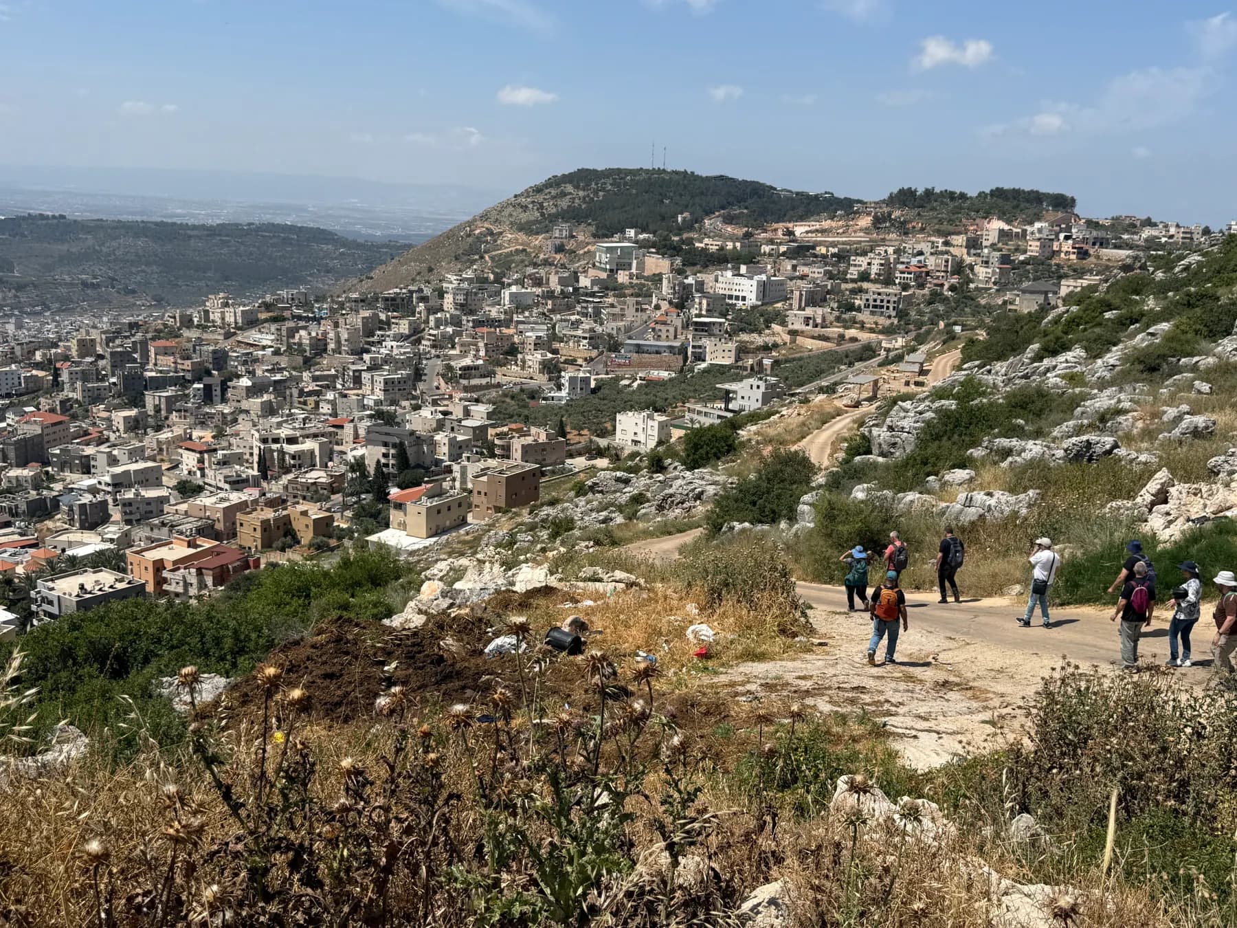 Houses of Deir al-Asad spreading over a hillside, seen from the Zurim cliff, with hikers on a road below.