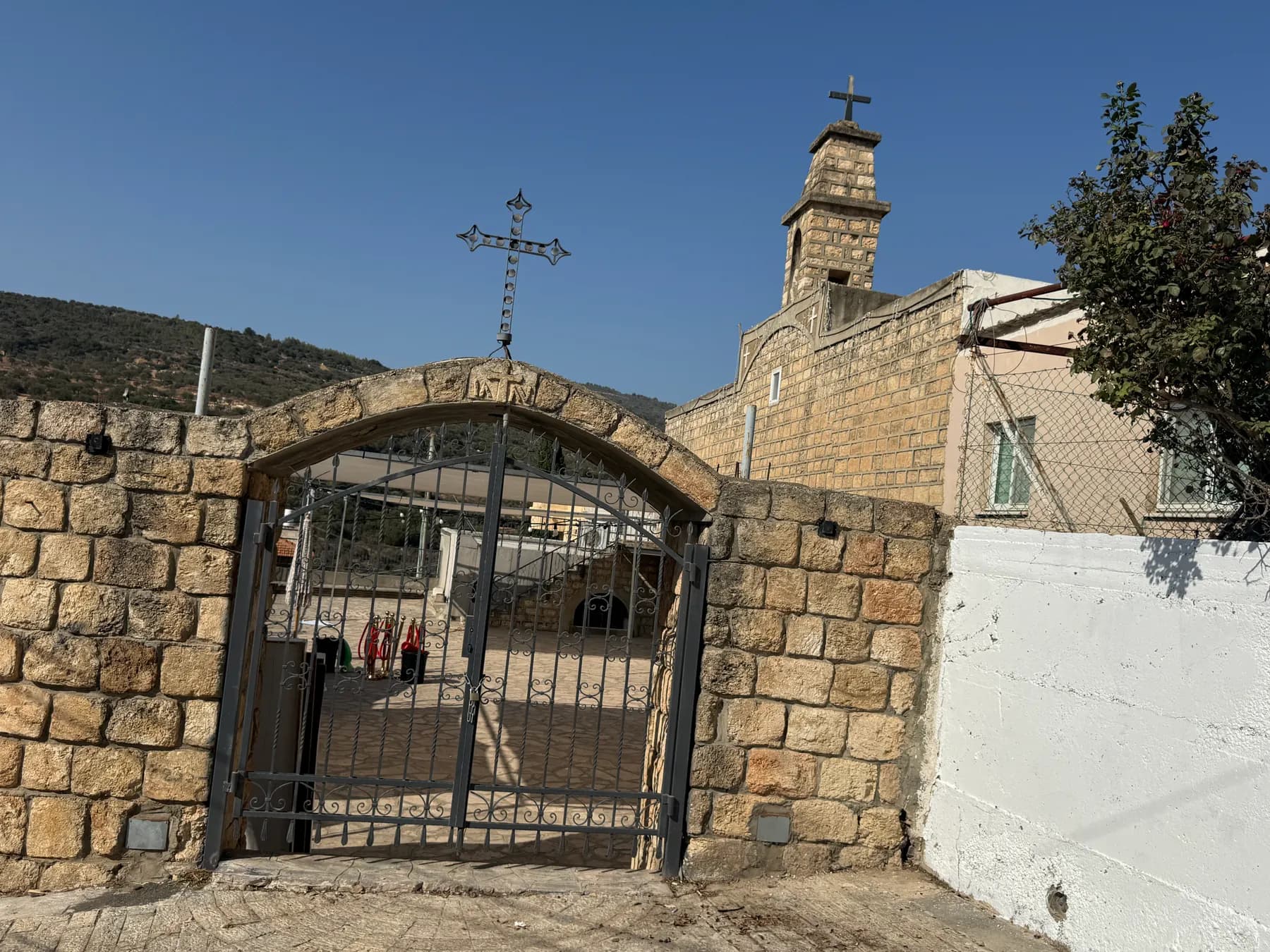 An arched stone gateway with an iron gate and a cross leading to an old stone church with a bell tower in Eilabun.